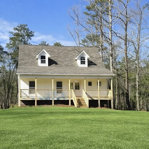 A yellow custom farmhouse with a gray roof and white porch, surrounded by green grass and trees under a blue sky.