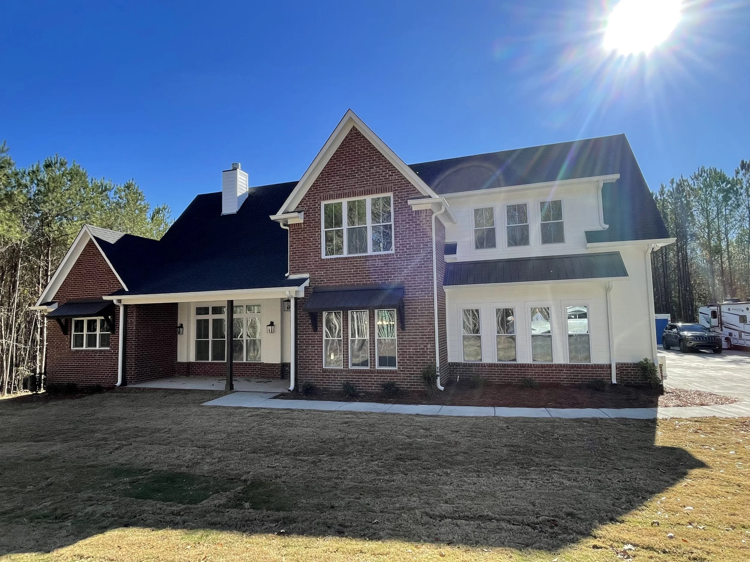 Newly constructed custom build two-story house with brick and white siding exterior, multiple large windows, black roof, and a small front porch, under bright sunlight with a clear blue sky and trees in the background.