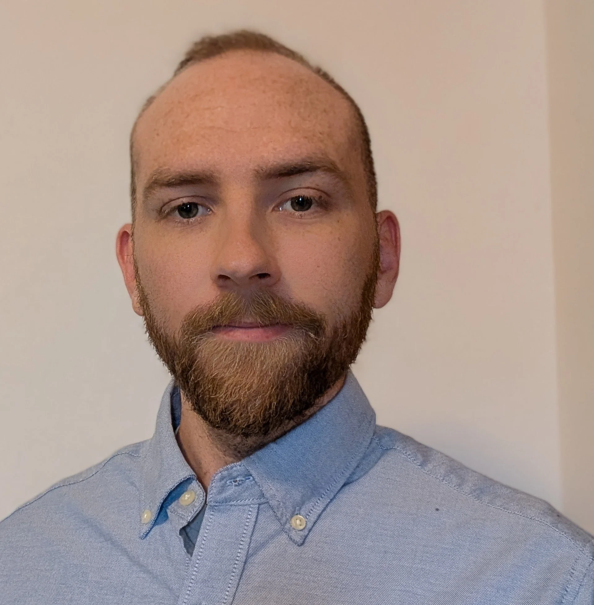 Close-up of a man with a reddish beard and mustache, wearing a light blue collared shirt, standing against a plain light-colored wall.