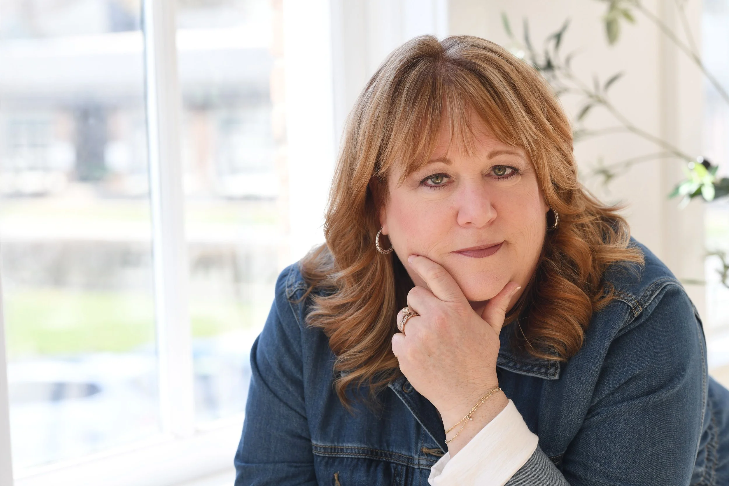 A woman with reddish-brown hair and green eyes resting her chin on her hand, located near a window with blurred outdoor scenery in the background.