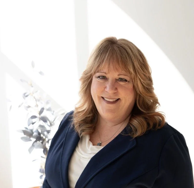 A middle-aged woman with shoulder-length reddish-blonde hair, smiling, wearing a navy blazer and a white top, standing indoors near a window with natural light.