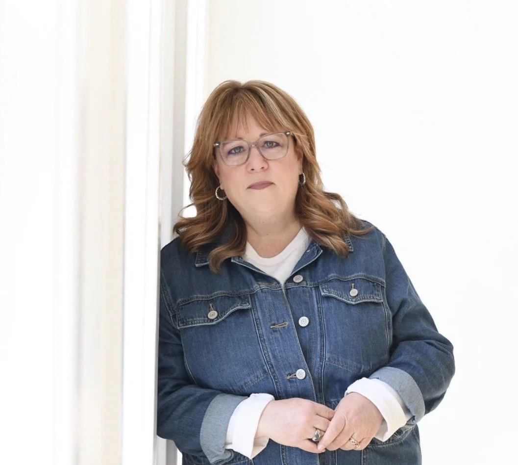 A woman with reddish-brown hair and glasses wearing a denim jacket over a white shirt, standing against a white background.