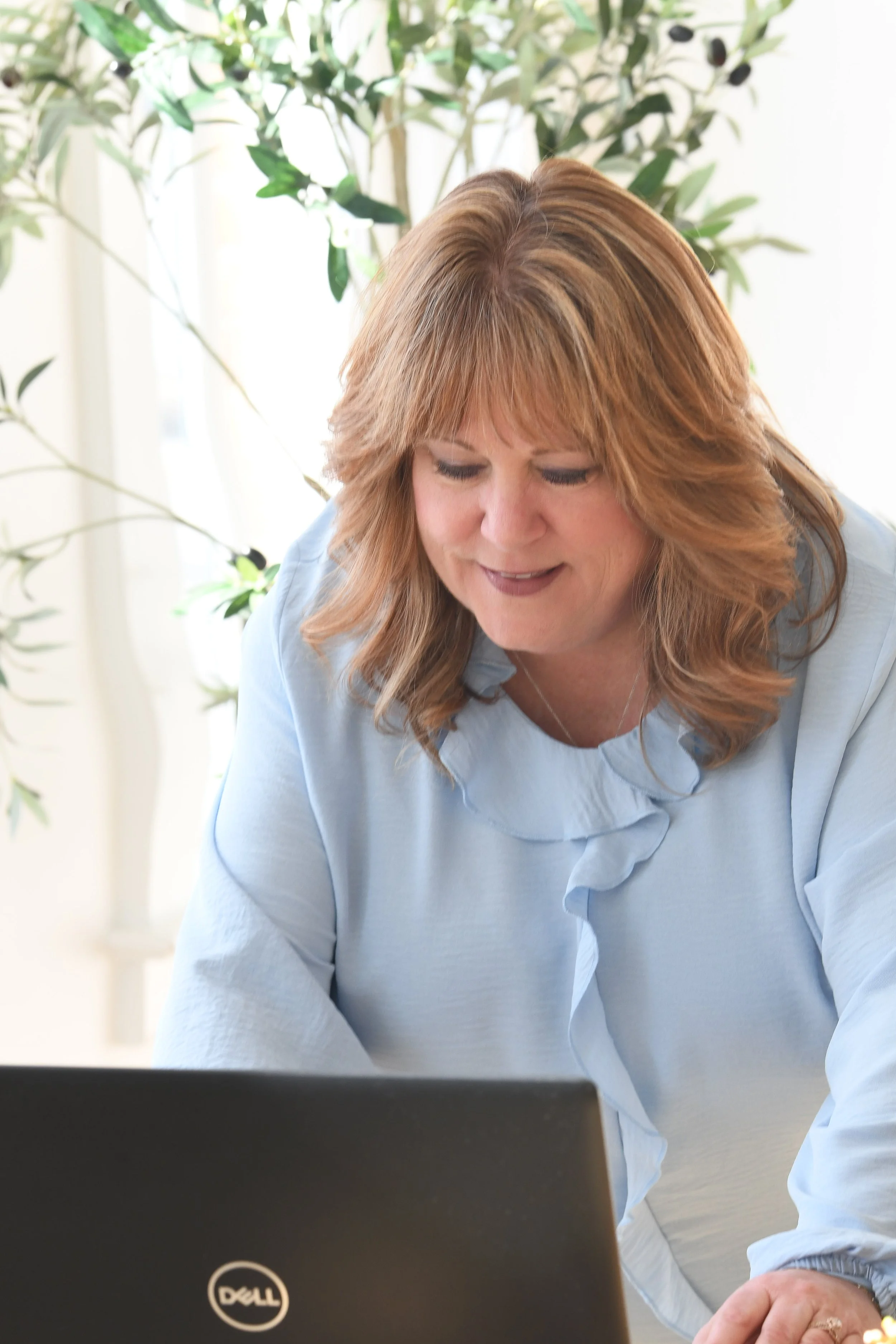 Woman with reddish-brown hair working on a laptop in a bright room with a large green plant in the background.