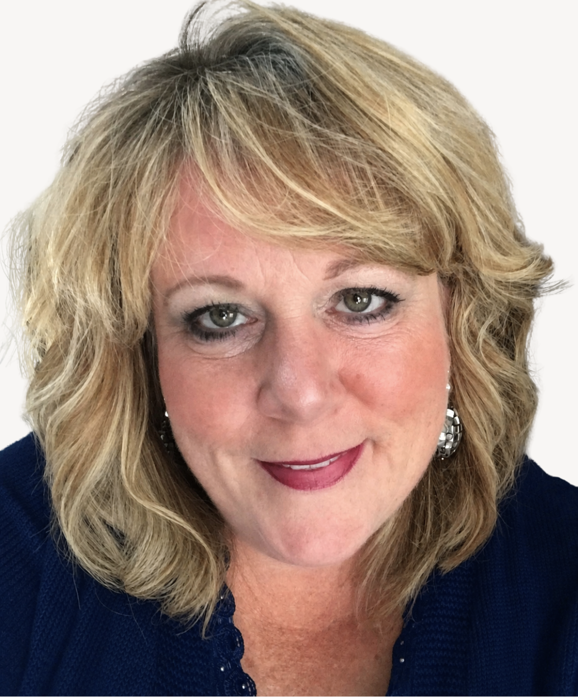 A close-up portrait of a middle-aged woman with shoulder-length curly blonde hair, green eyes, and wearing silver earrings, a dark blue top, and makeup including lipstick and eyeshadow, smiling softly against a plain white background.