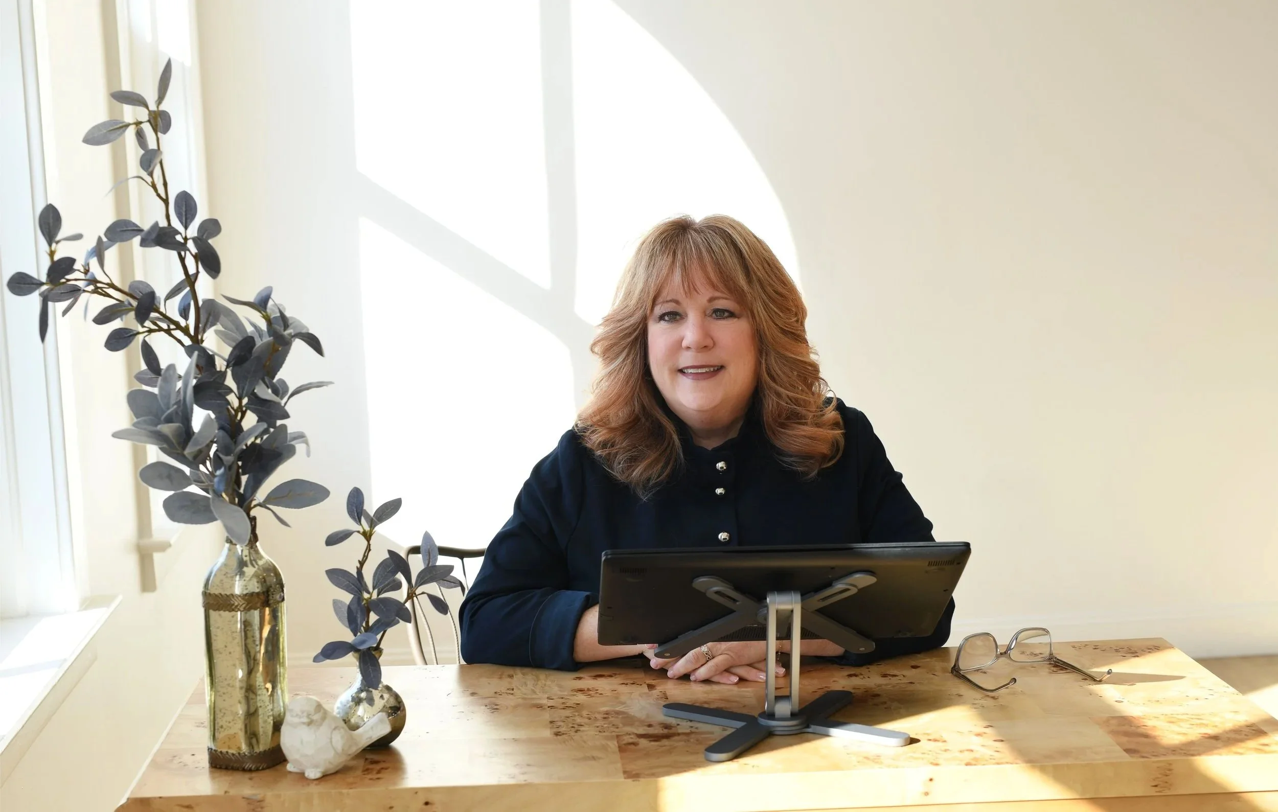 A woman with red hair and a black top sitting at a wooden table, with a laptop in front of her. To her left, decorative vases with dark green leaves. She is indoors near a window with sunlight casting shadows.
