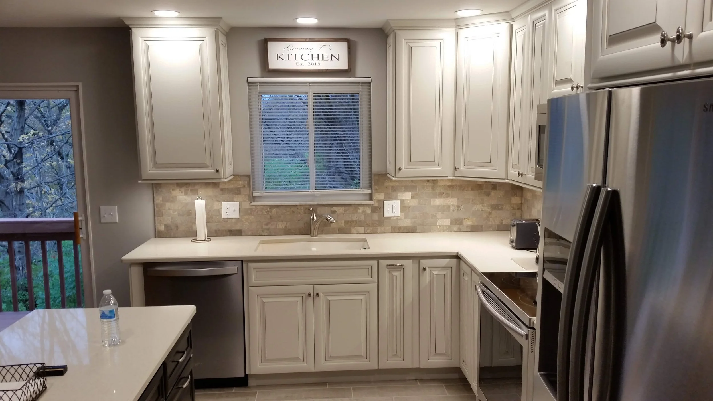 Kitchen with white cabinets, a window with blinds, a beige tile backsplash, stainless steel refrigerator, stove, and sink, with a sign above the window that says 'Grammy T's Kitchen Est. 2018'.