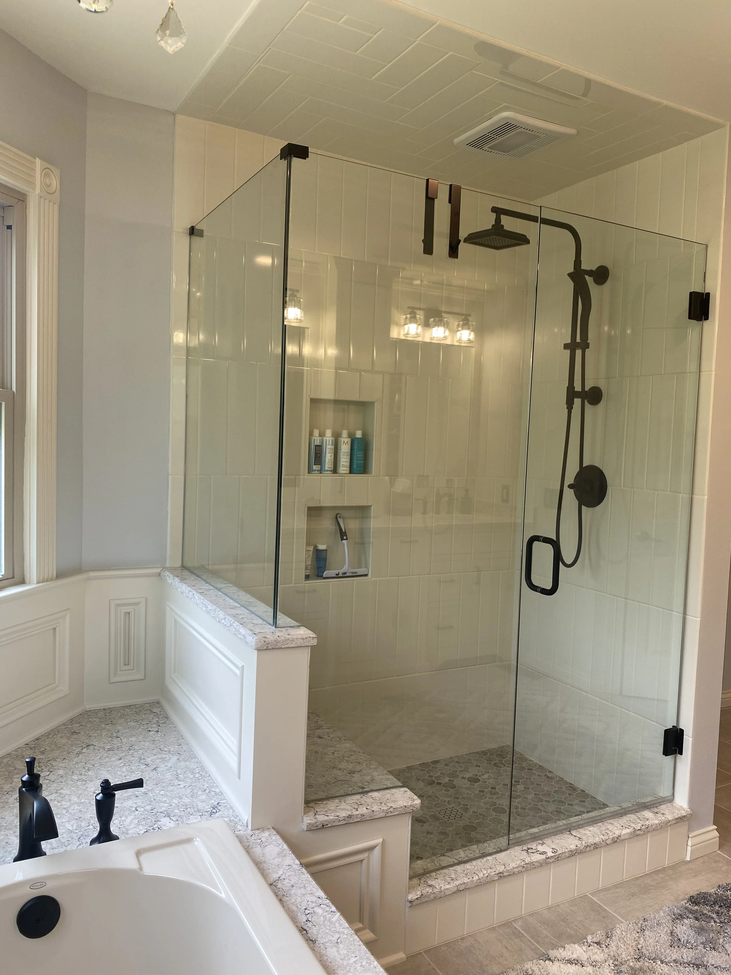 A glass-enclosed bathroom shower with beige tiles, a black shower system, built-in shelves with toiletries, and a view of a bathtub with black fixtures nearby.