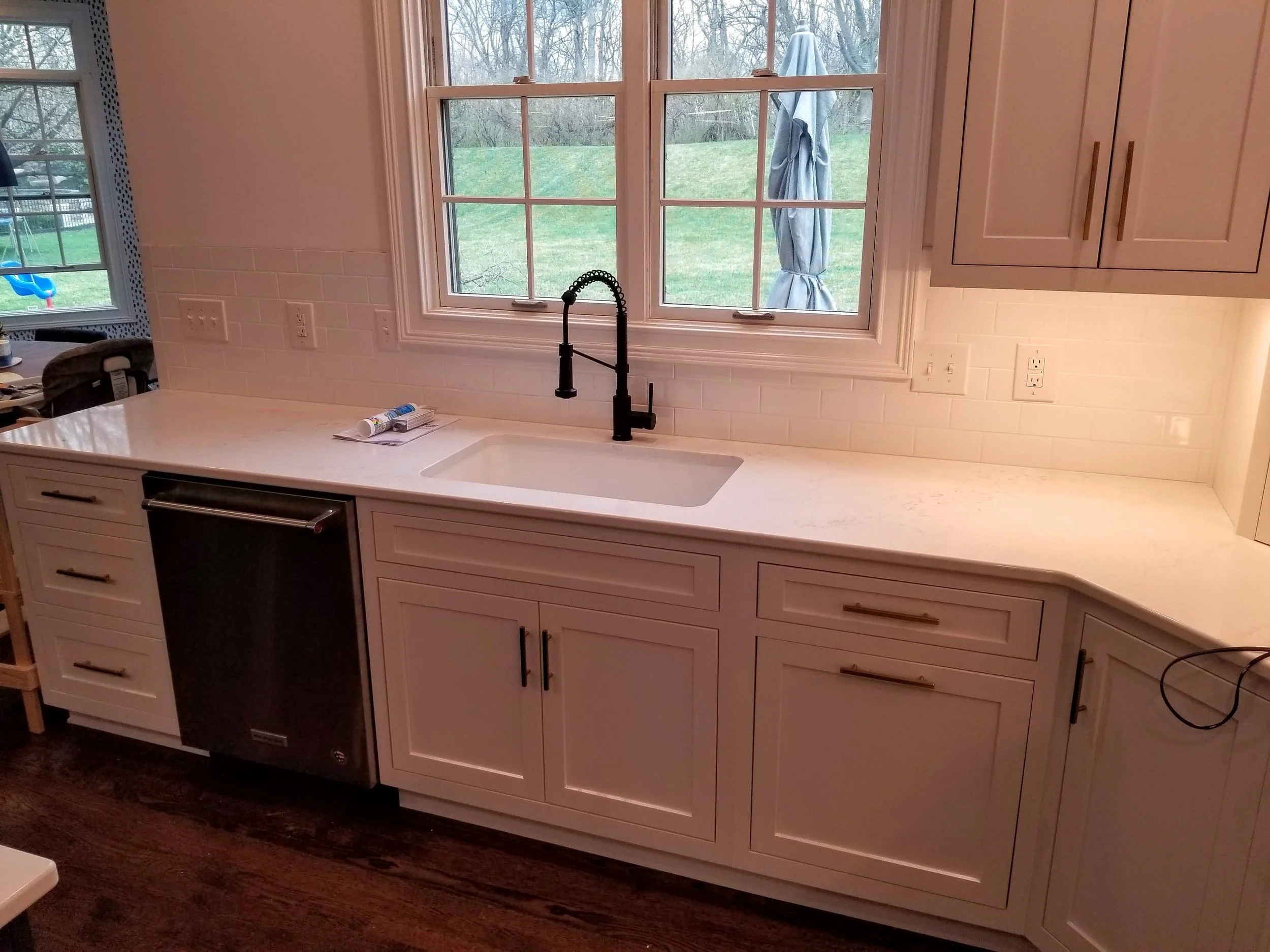 A kitchen countertop with a black faucet, under two large windows overlooking a grassy yard with trees. There is a small built-in dishwasher beneath the counter and beige cabinets above and below, with a light-colored backsplash and a dark wood floor