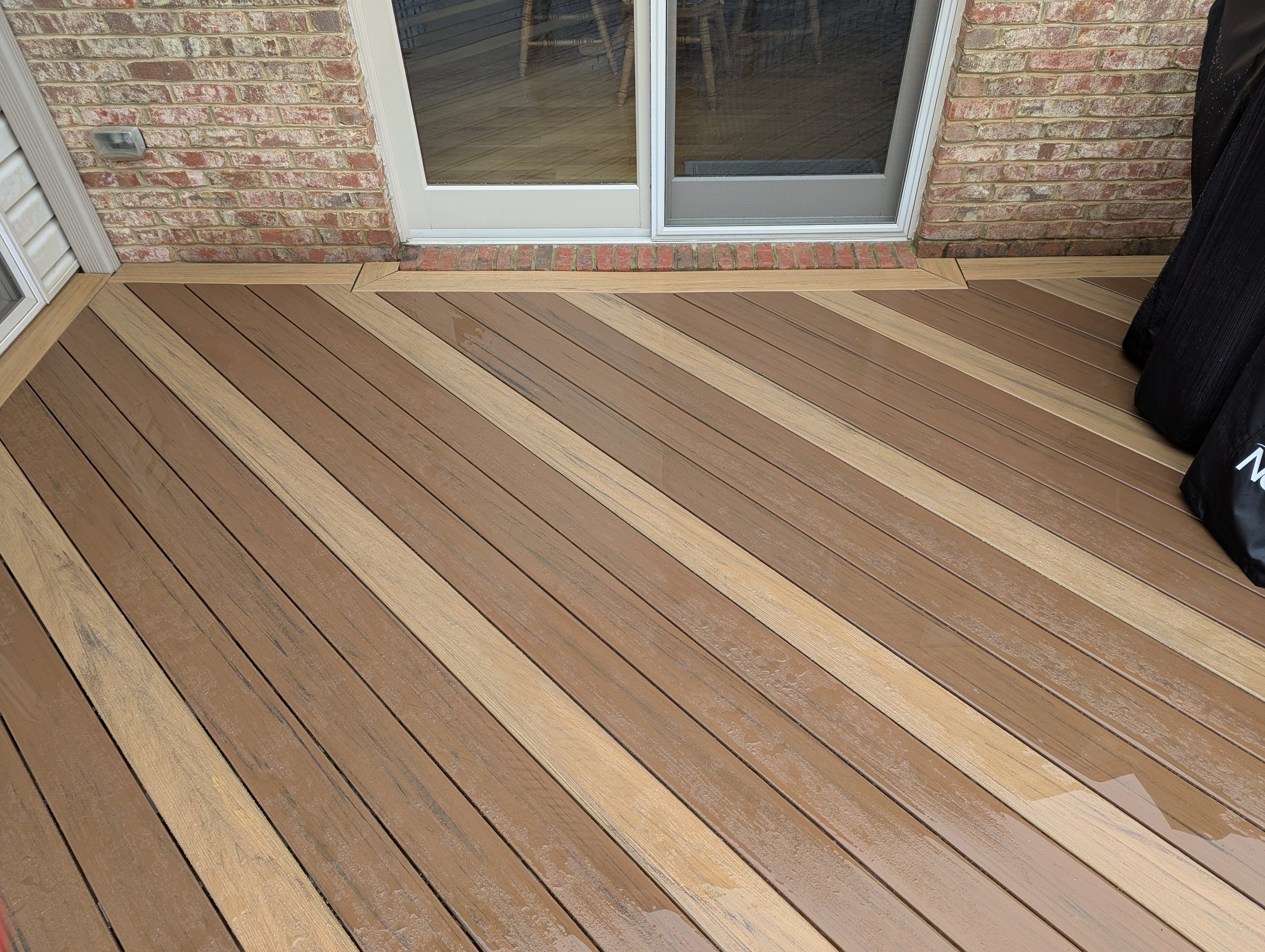 A newly installed wooden deck with alternating light and dark brown planks outside a house, adjacent to sliding glass doors with brick exterior walls.