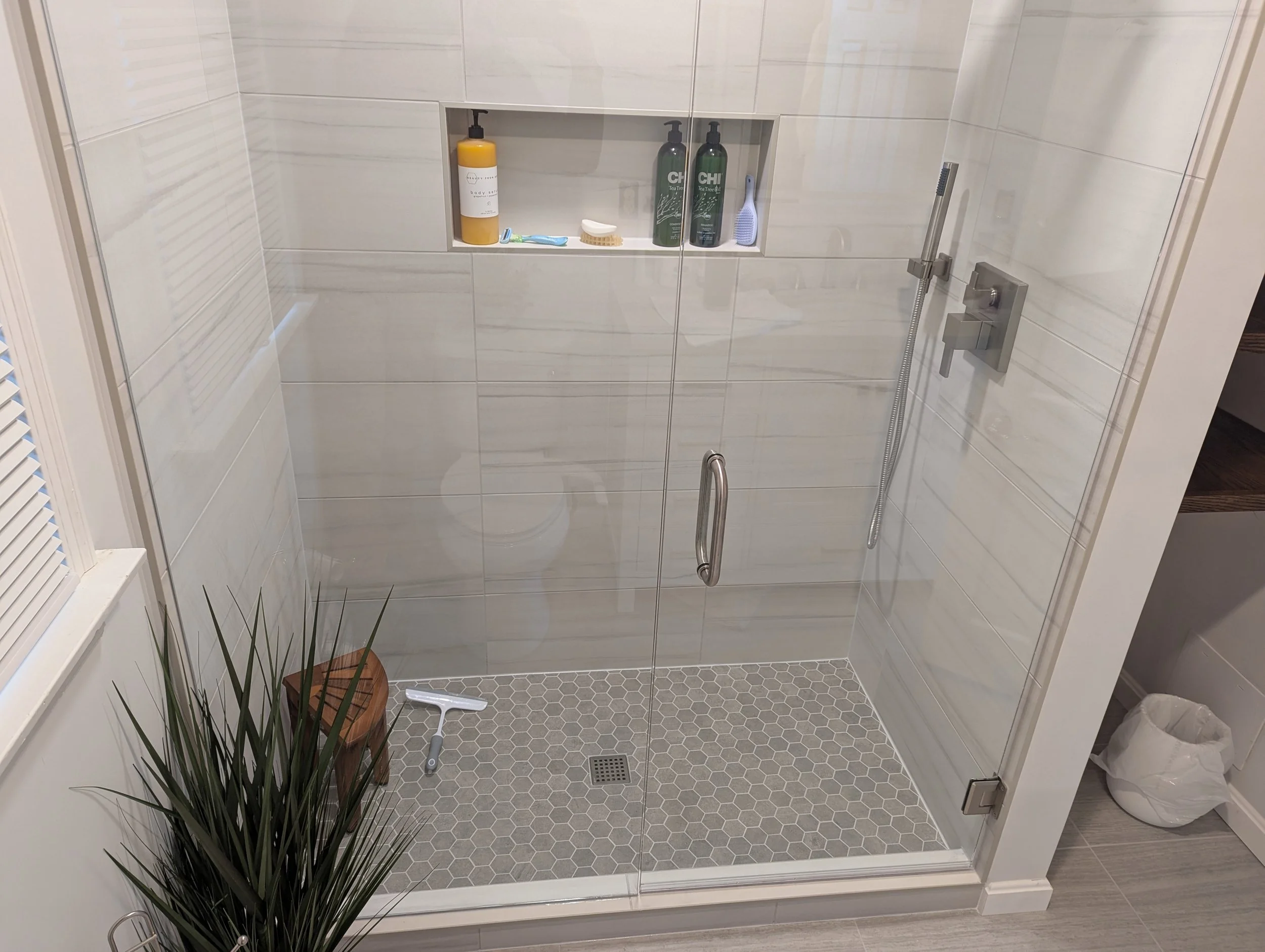 Modern glass shower enclosure with tiled walls and hexagon tile floor, interior recessed shelf with toiletries, a cleaning squeegee on the floor, a potted plant outside, and a trash can nearby.