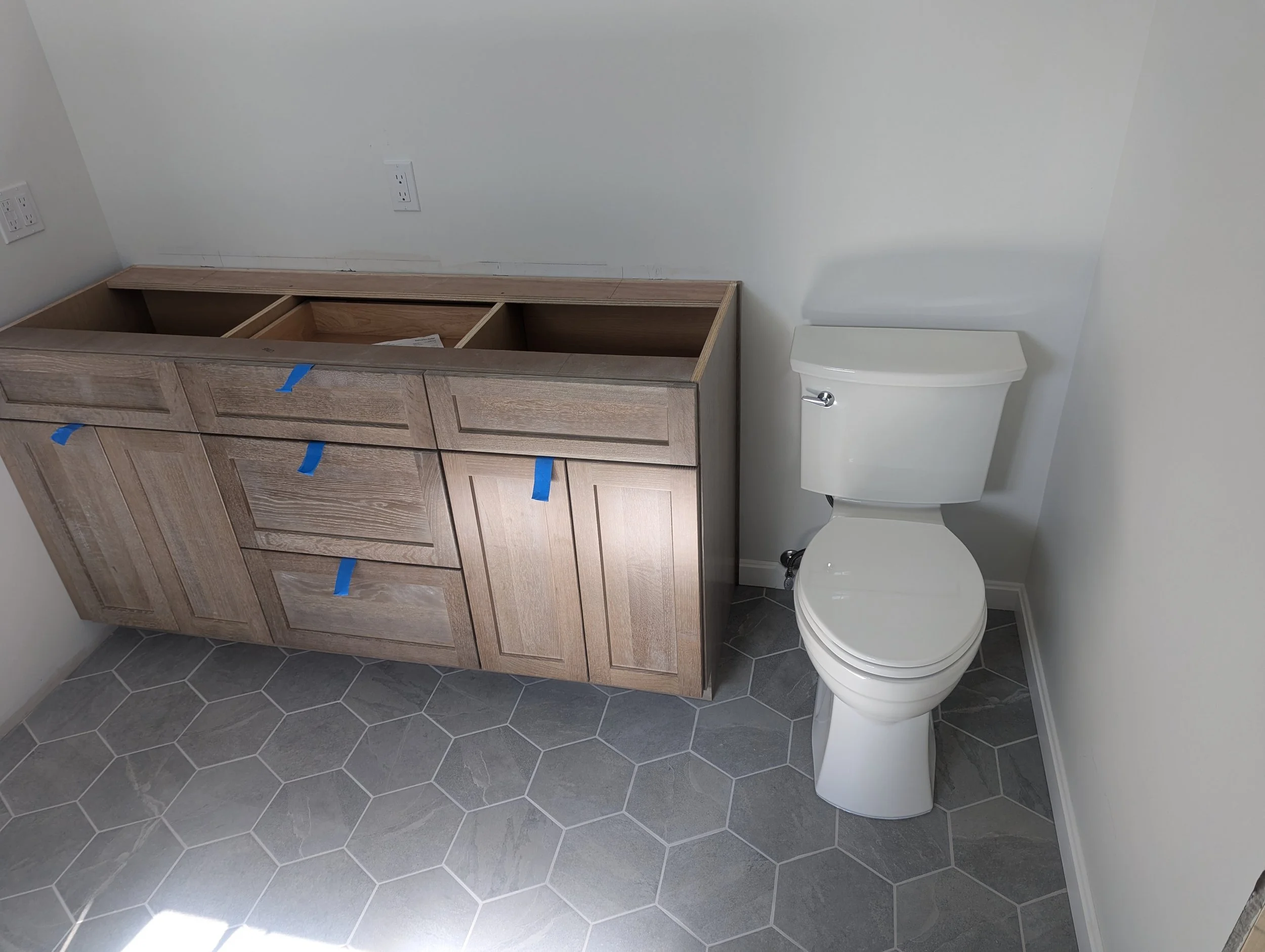 Bathroom with a vanity cabinet under construction and a white toilet, gray hexagonal tile flooring, and a plain white wall.