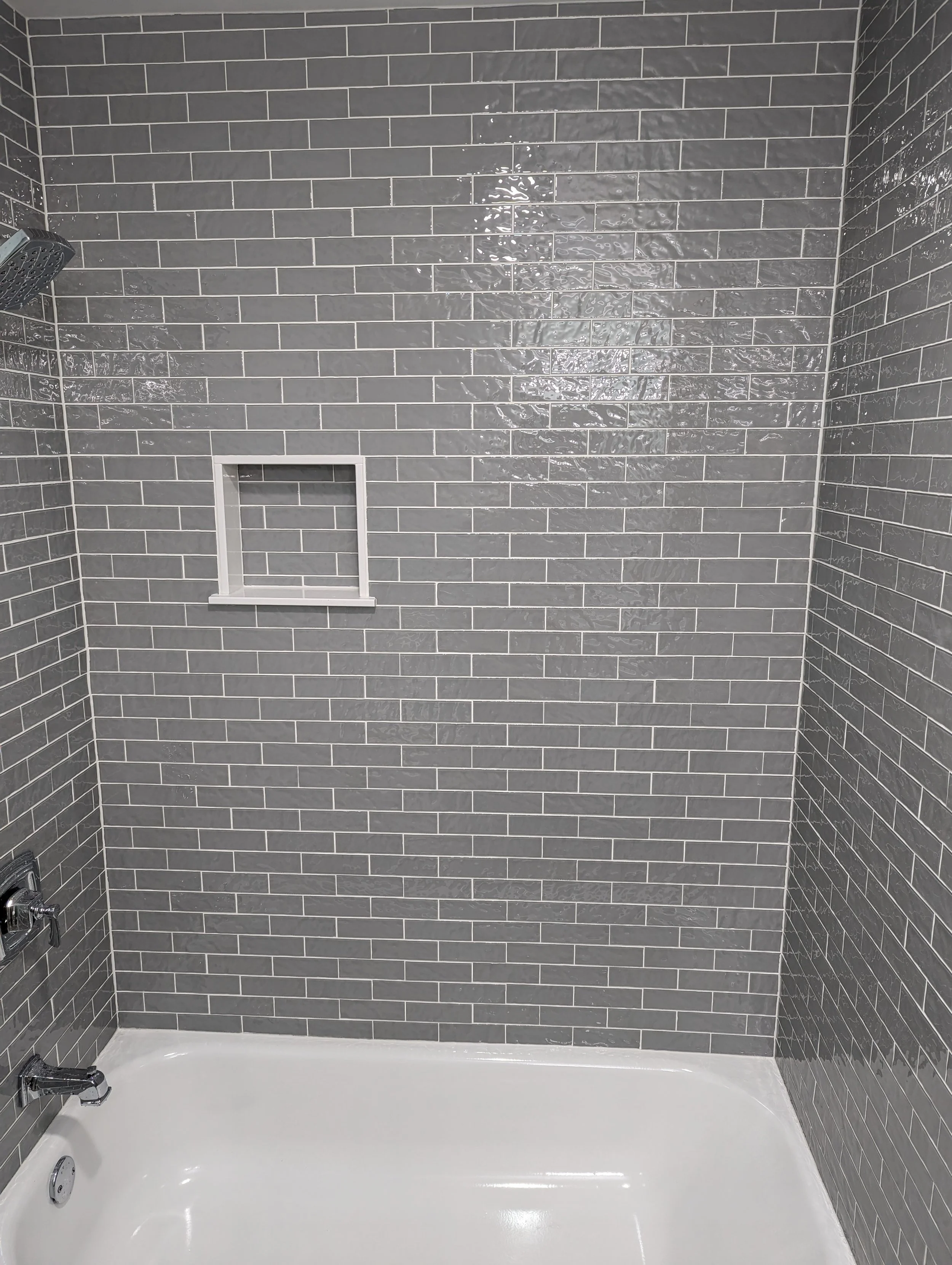 A shower stall with gray subway tile walls, a built-in niche, a showerhead, and a bathtub.