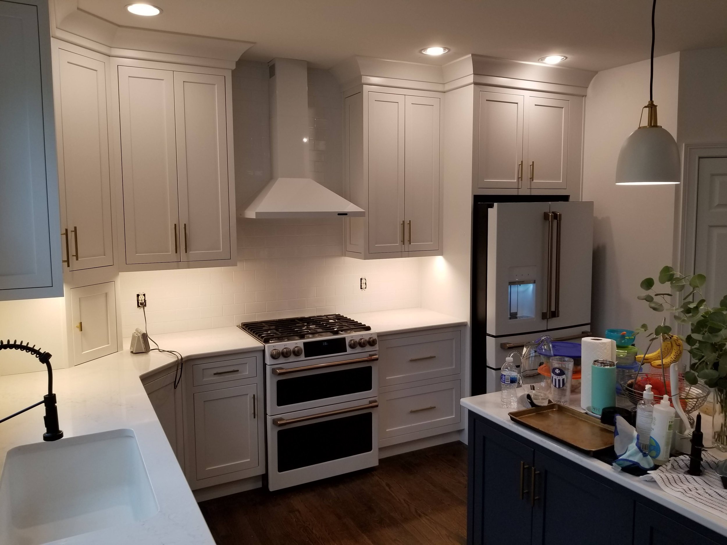 Modern kitchen with white cabinets, a white range hood, a double oven, a refrigerator, and a kitchen island with various items and kitchen essentials.