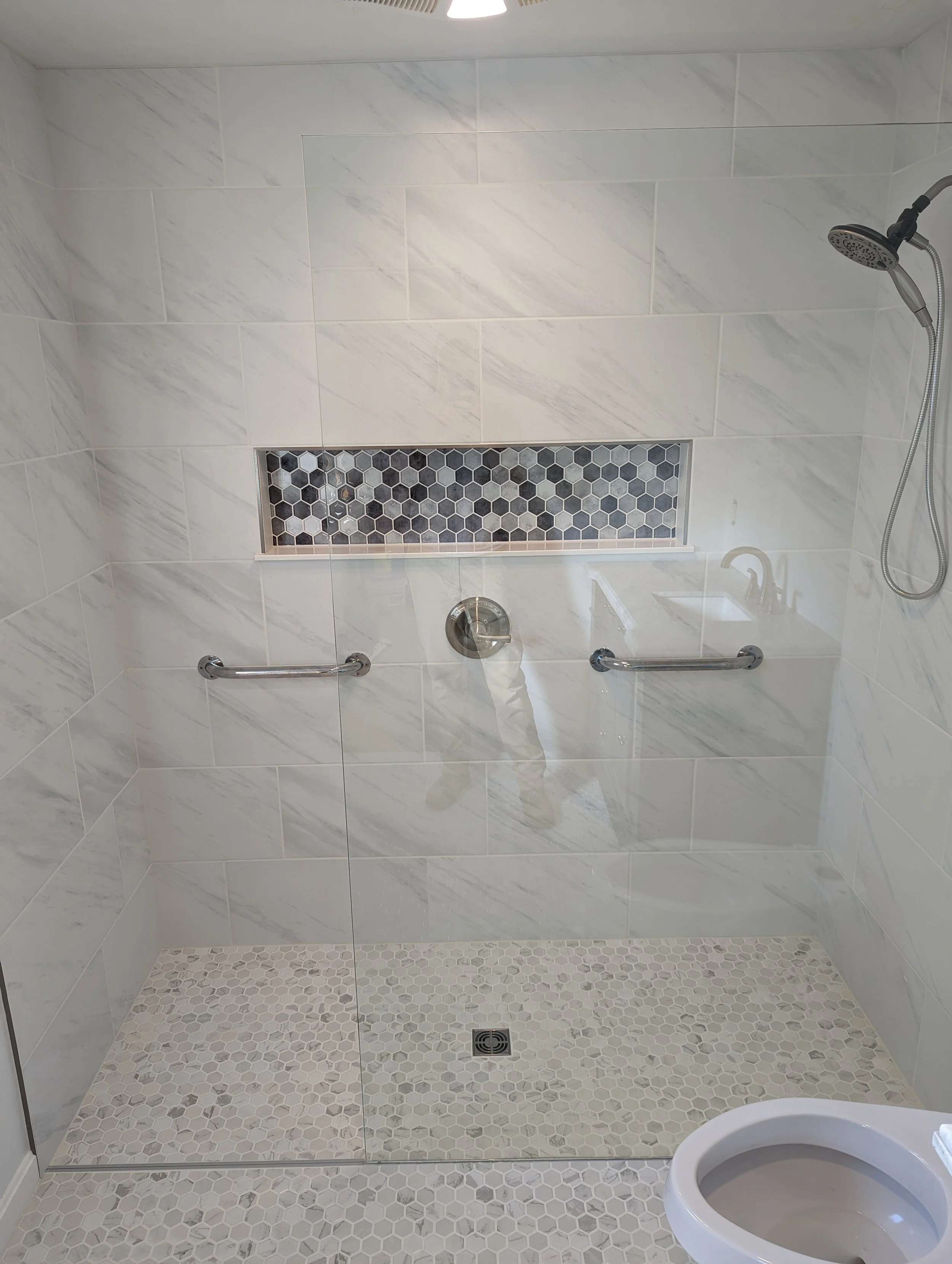 Accessible walk-in shower with marble tile walls, a hexagonal tile floor, a built-in niche with small hexagonal tiles, grab bars, a handheld showerhead, and a drain in the floor.