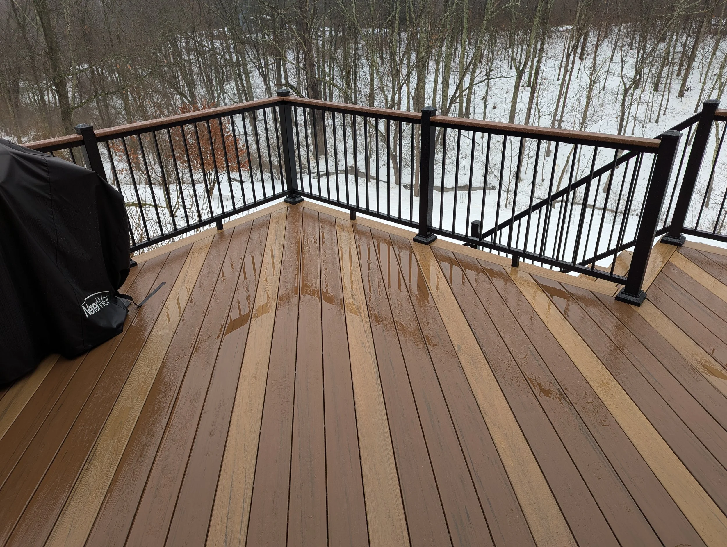 A wooden deck with wet surface, black metal railing, and a covered grill on the left, overlooking a snowy landscape of trees in the background.
