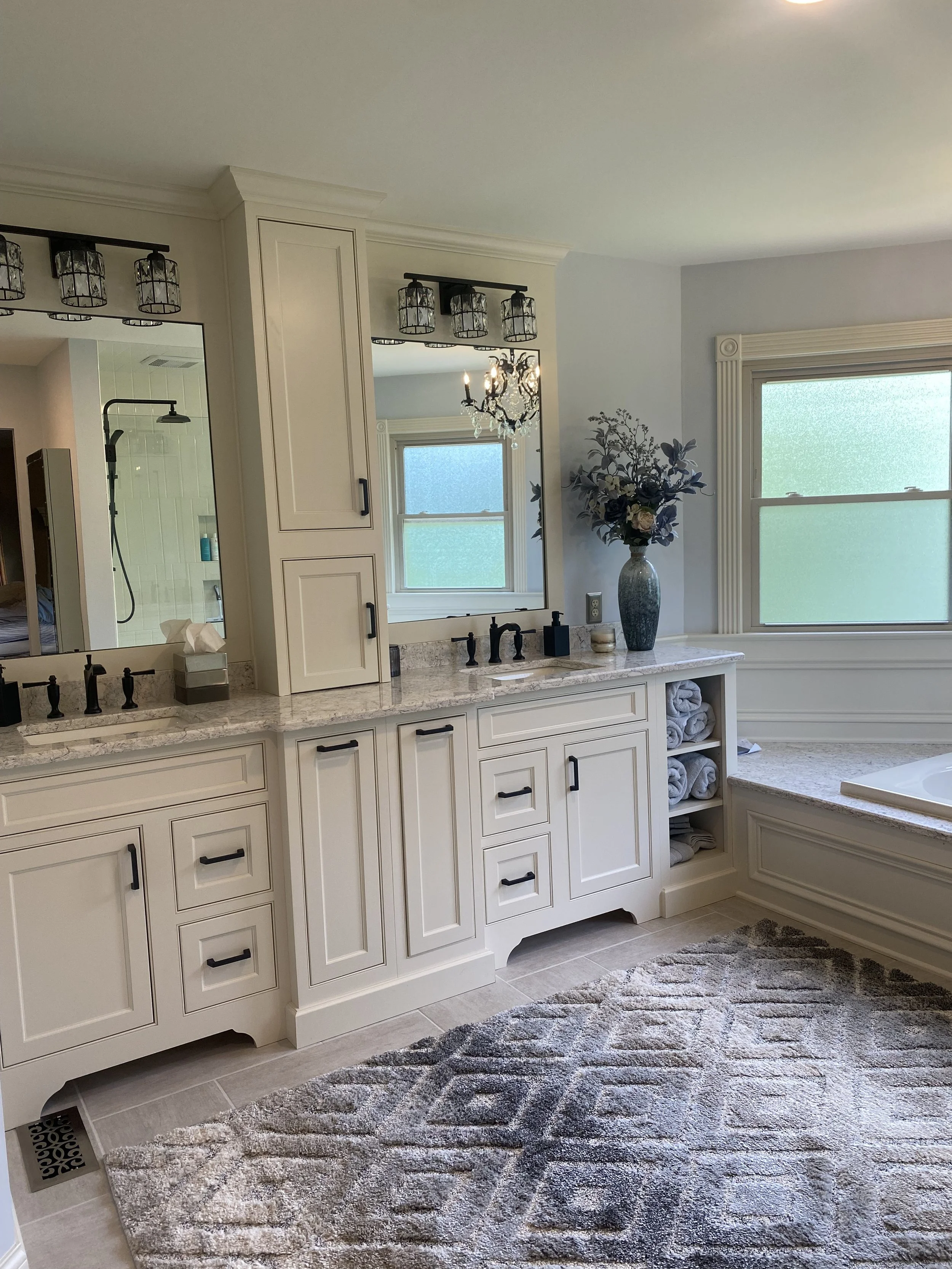 A spacious bathroom with a large vanity featuring a granite countertop, black fixtures, and white cabinetry. A mirror with vintage-style light fixtures is above the vanity. There is a window with frosted glass, a decorative vase with flowers, rolled 