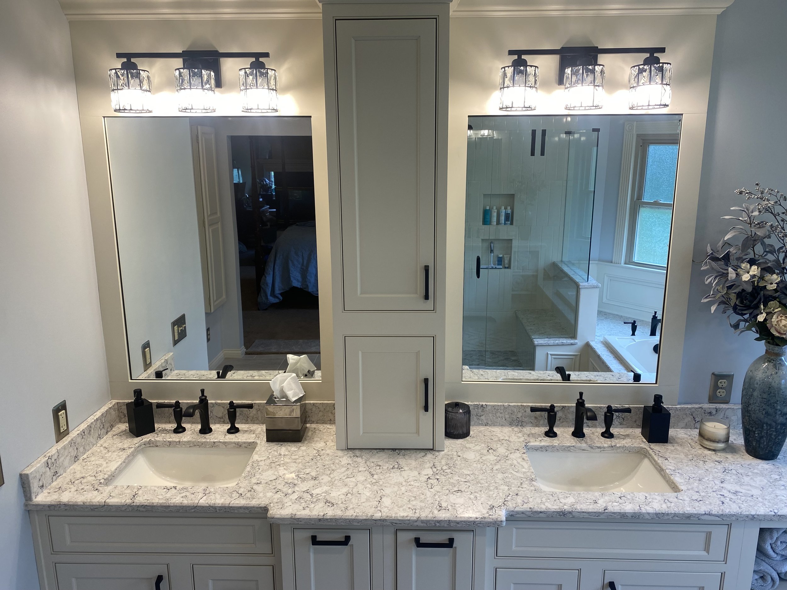 A bathroom with a double vanity featuring marble countertops, two sinks, and two large mirror panels. The left side has tissue box and soap dispenser, the right side has a candle and decorative vase with flowers. Overhead lighting with two fixtures e