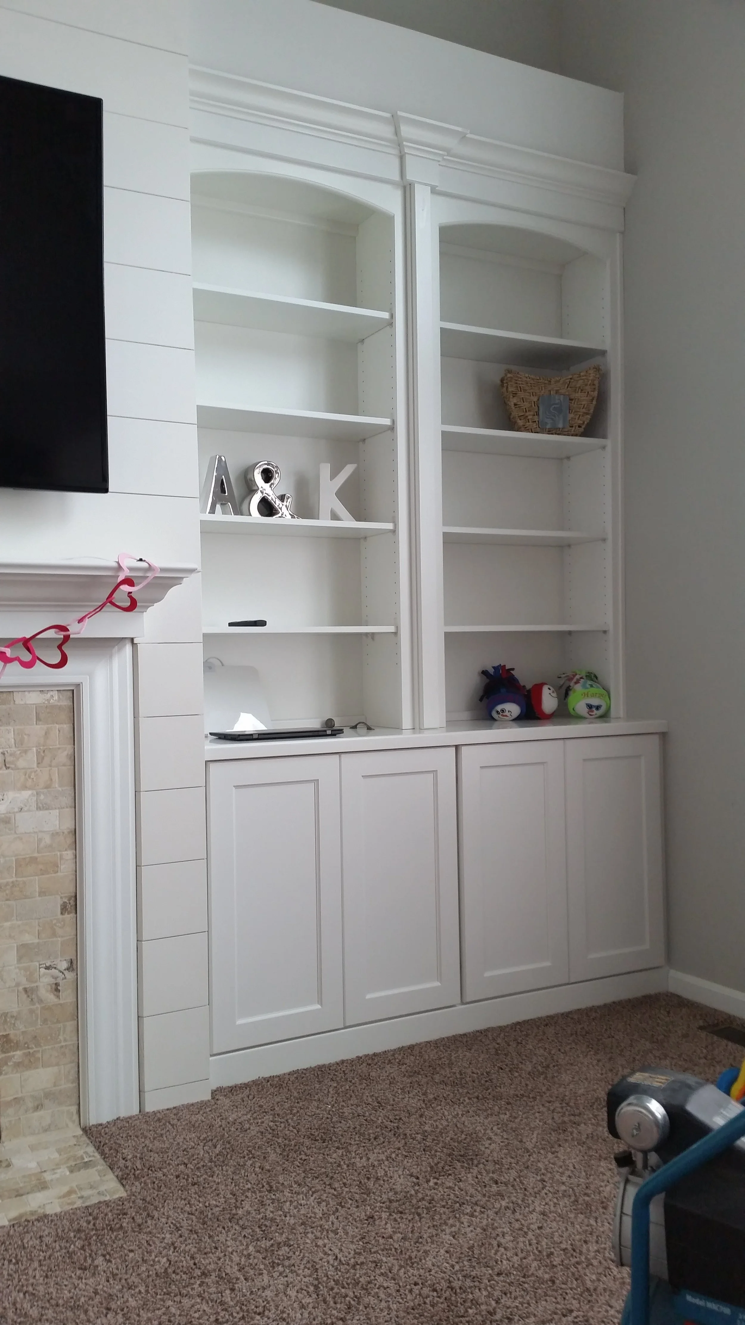 White built-in bookshelf with three shelves, some decorative letters, stuffed toys, and a small wicker basket, adjacent to a fireplace with a pink heart garland, in a living room.