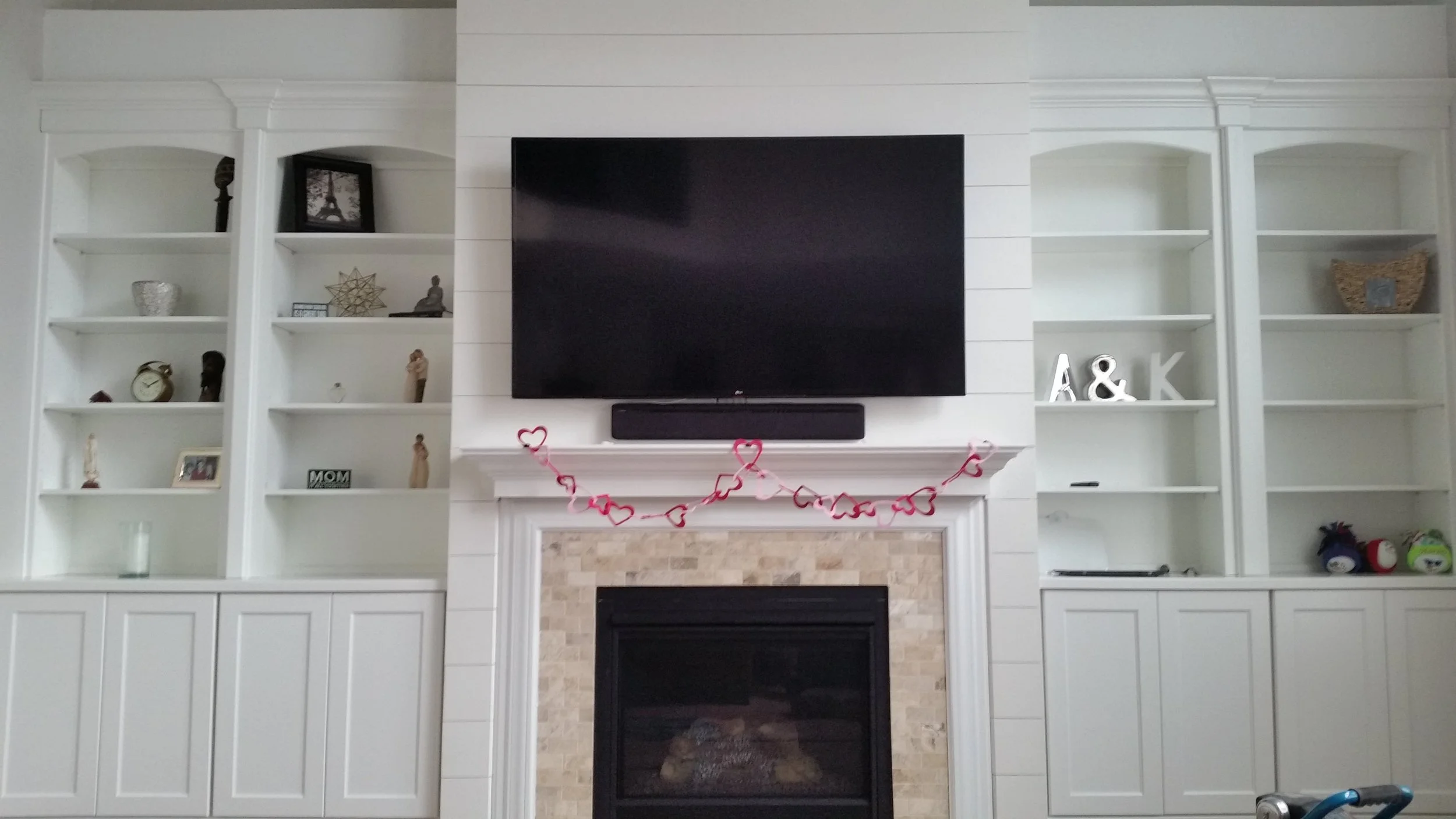Living room entertainment center with a mounted flat-screen TV, a pink heart garland, built-in white bookshelves on either side with decorative items, and a fireplace with a tile surround below the TV.