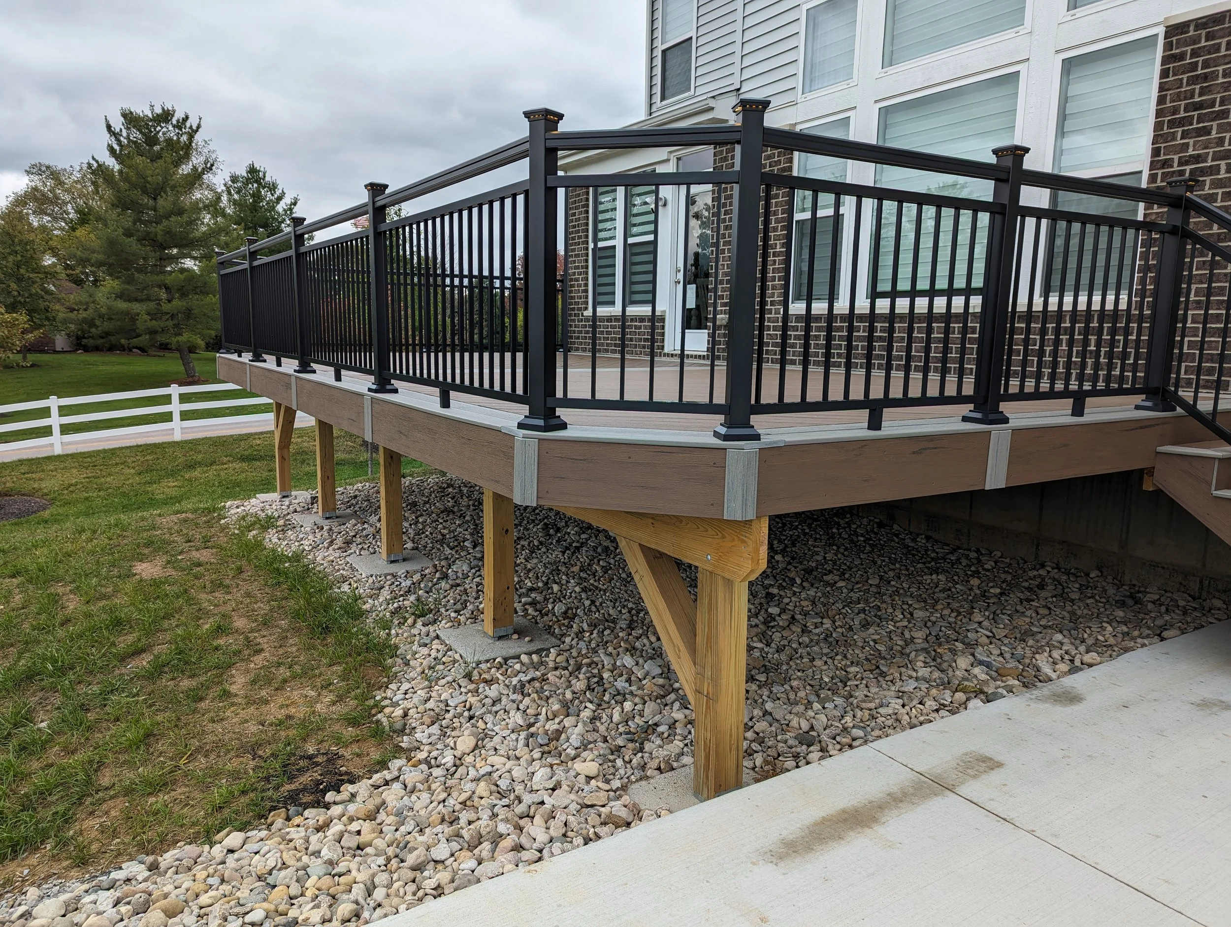 A newly built wooden deck with black metal railing attached to a brick house, supported by wooden posts and concrete footings, overlooking a backyard with grass, rocks, and a white fence and trees in the background.