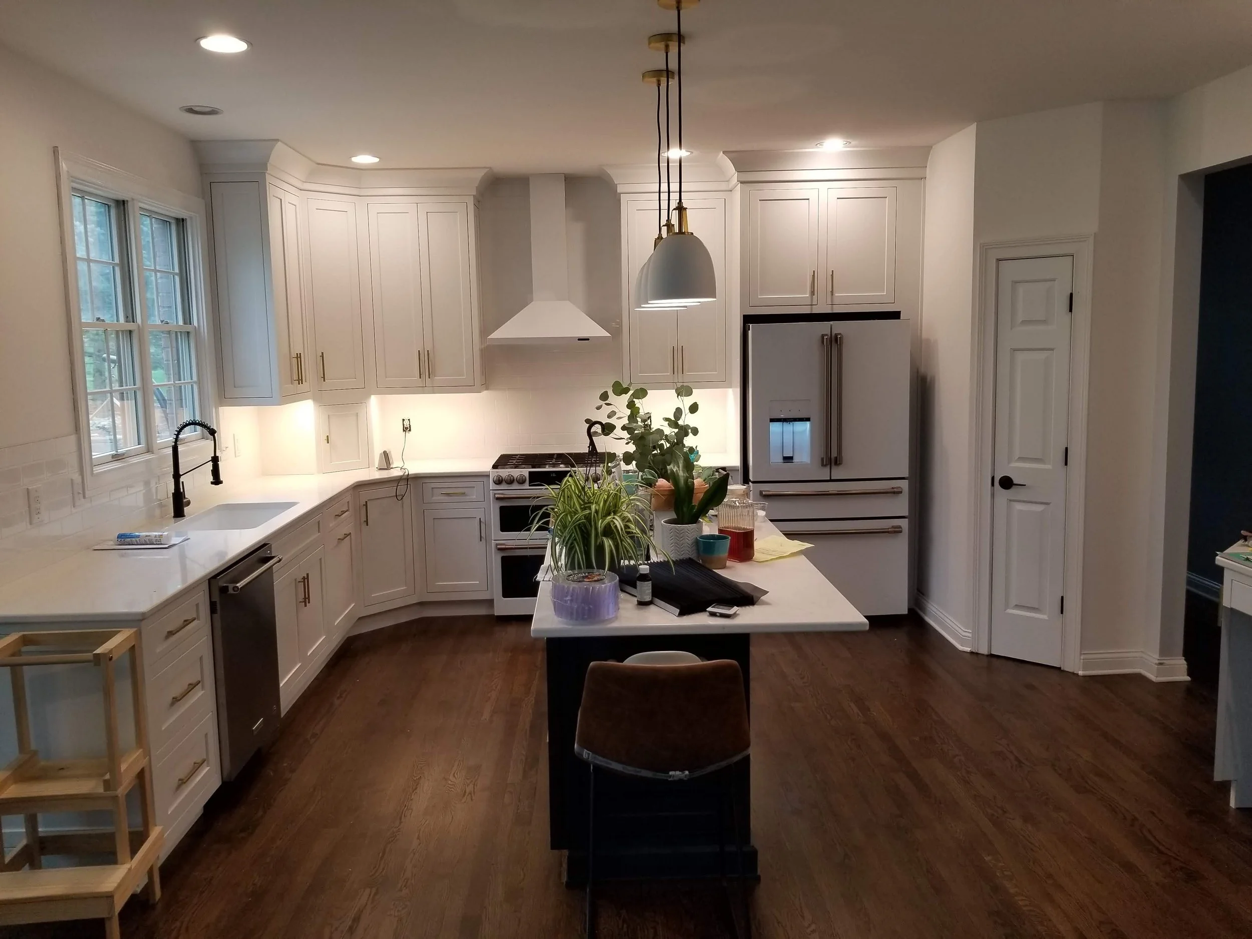 Modern white kitchen with hardwood floors, large windows, and a central island with plants and items on it, featuring upper and lower cabinetry, a white range hood, and a refrigerator.