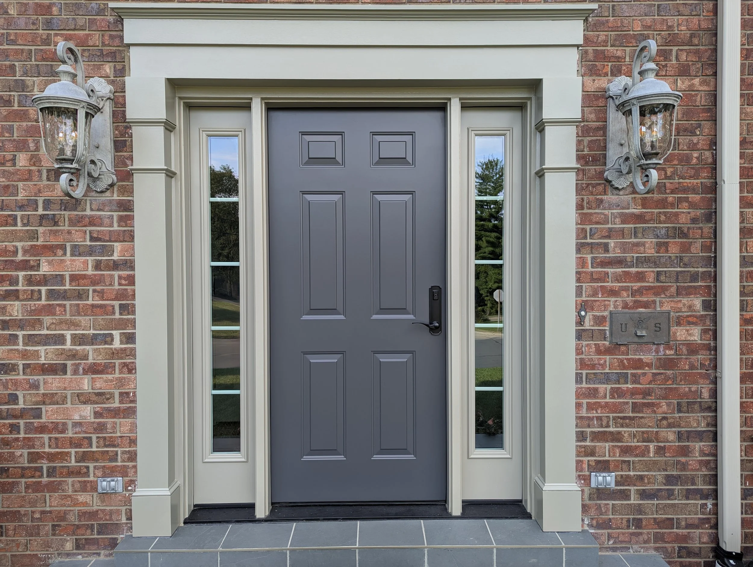 Front door of a brick house with a dark gray door, two vertical windows on either side, and two wall-mounted lanterns.