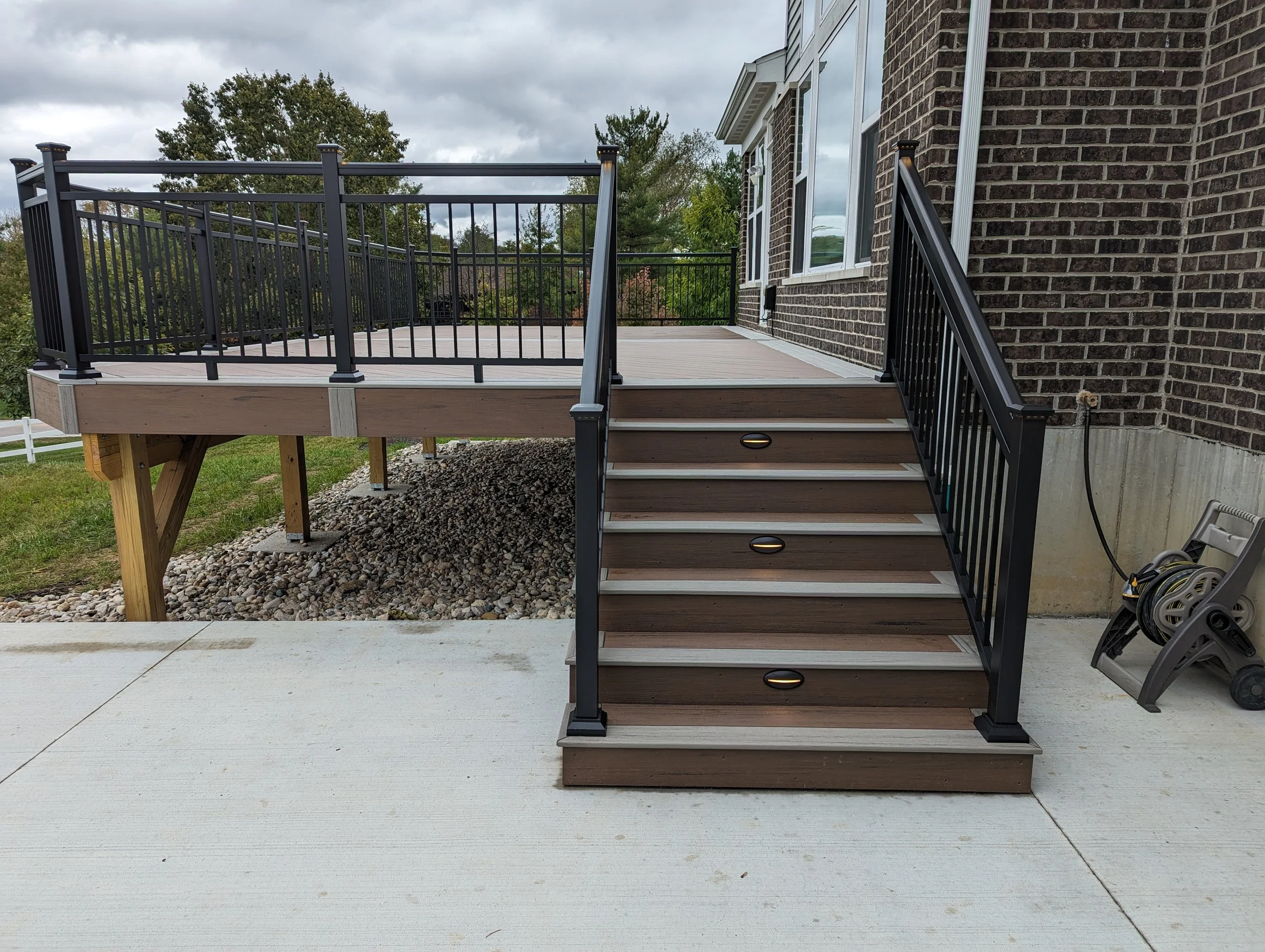 A newly built wooden deck with black metal railings, stairs, and a light fixture, attached to a brick house with a window, with landscaping and trees in the background.
