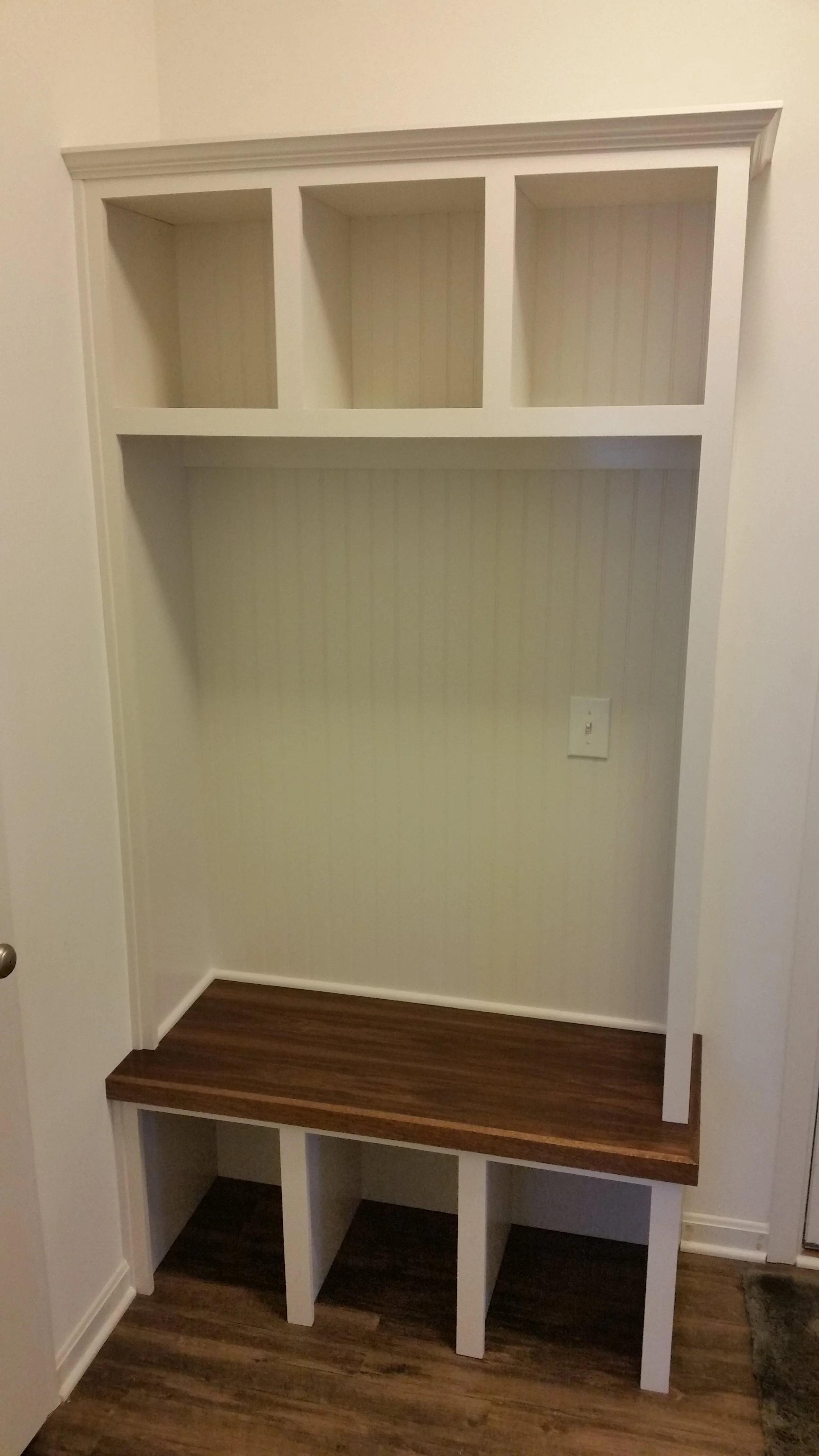 Empty built-in wall storage unit with three upper cubbies, a lower bench with a wood finish, and three open cubbies beneath, located in a room with wood flooring.
