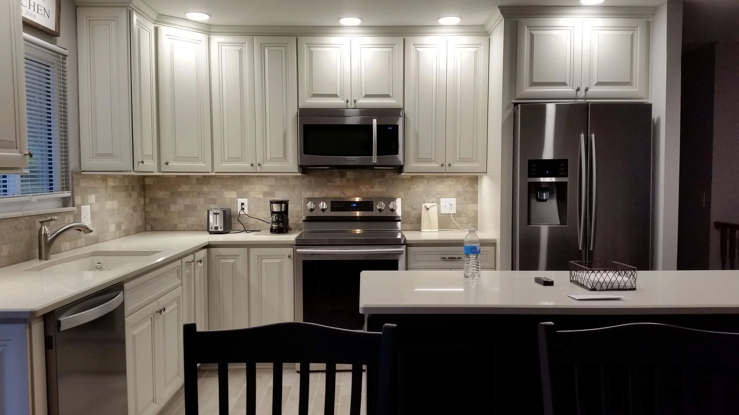 Kitchen with white cabinets, stainless steel appliances, coffee maker, toaster, water bottle, and black chairs at an island.