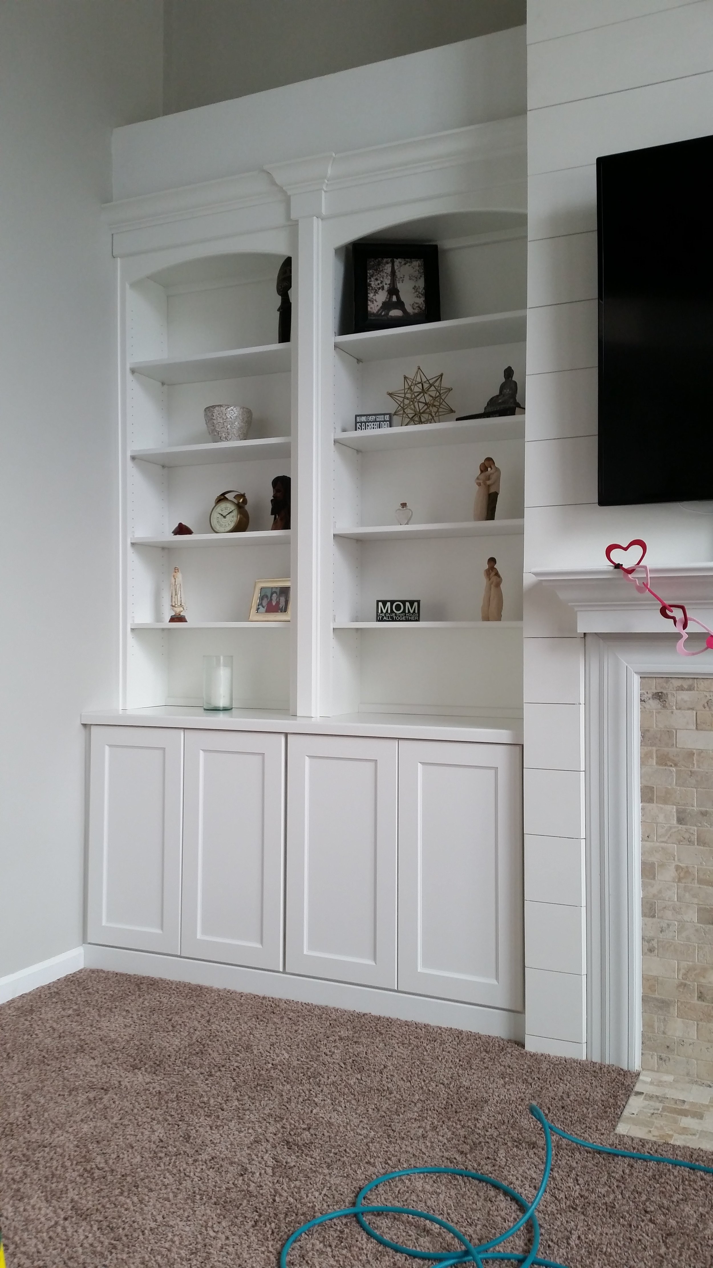 White built-in bookshelf with decorative items, a framed photo, and a small sign, situated next to a fireplace with a TV mounted above, in a living room with beige carpet.