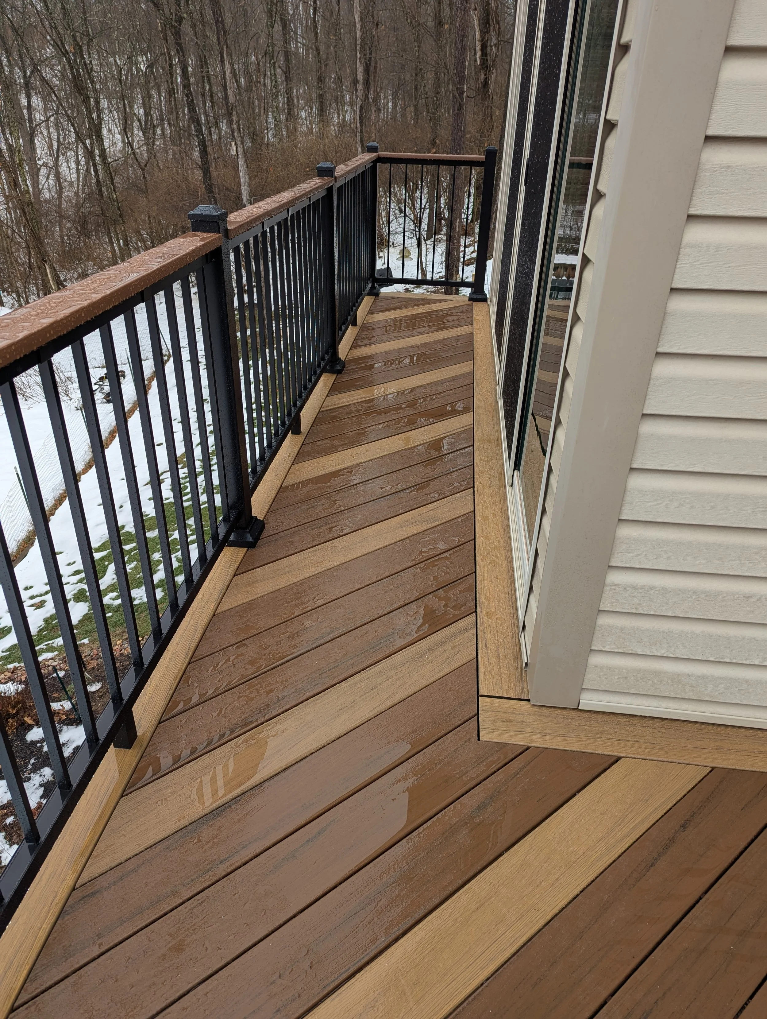 View of a wet outdoor balcony with wooden flooring and black metal railing, overlooking a snowy wooded area.