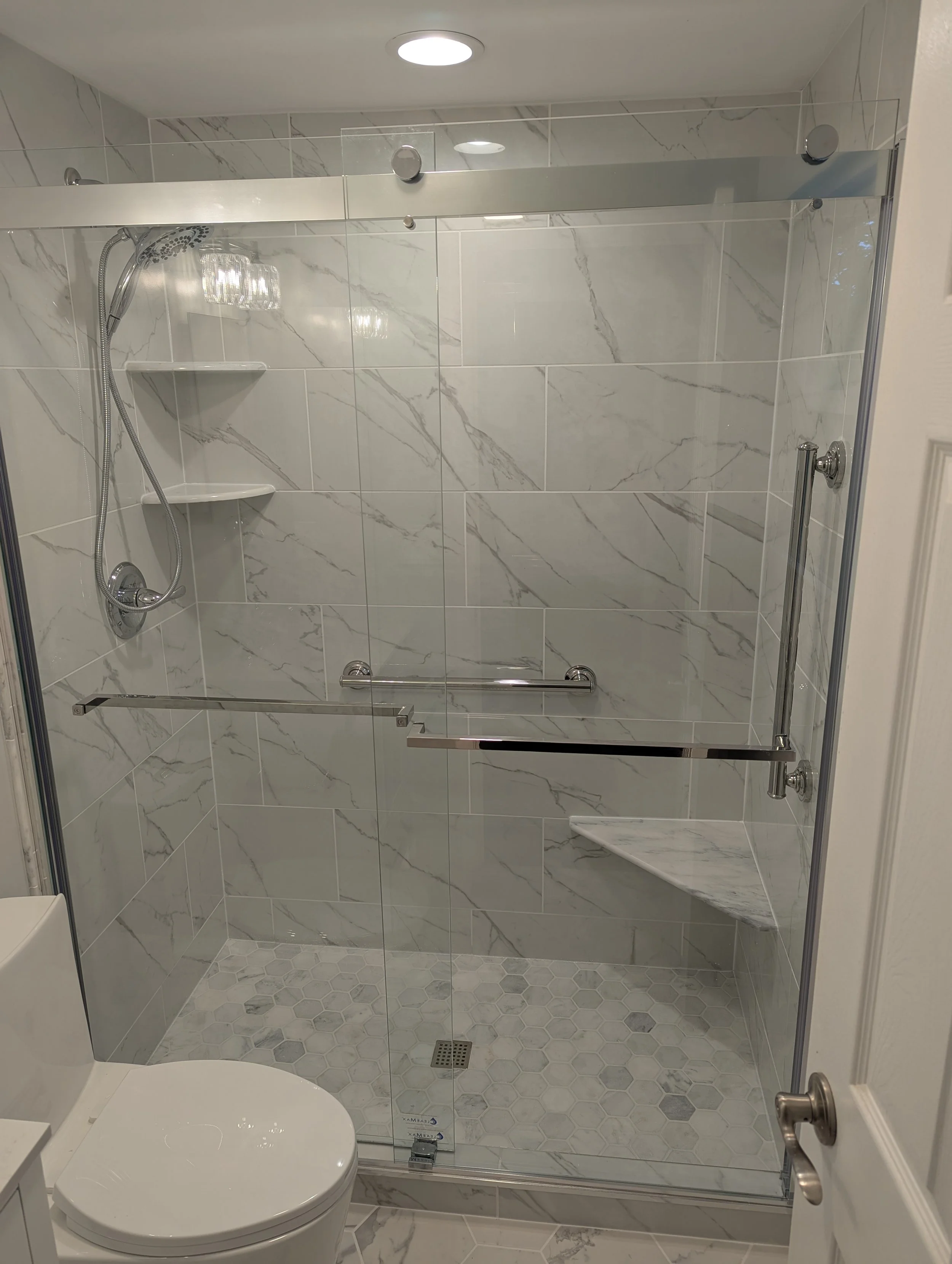 A modern shower with glass doors, marble tile walls, a handheld showerhead, built-in shelves, a grab bar, and a small corner marble bench, adjacent to a toilet.