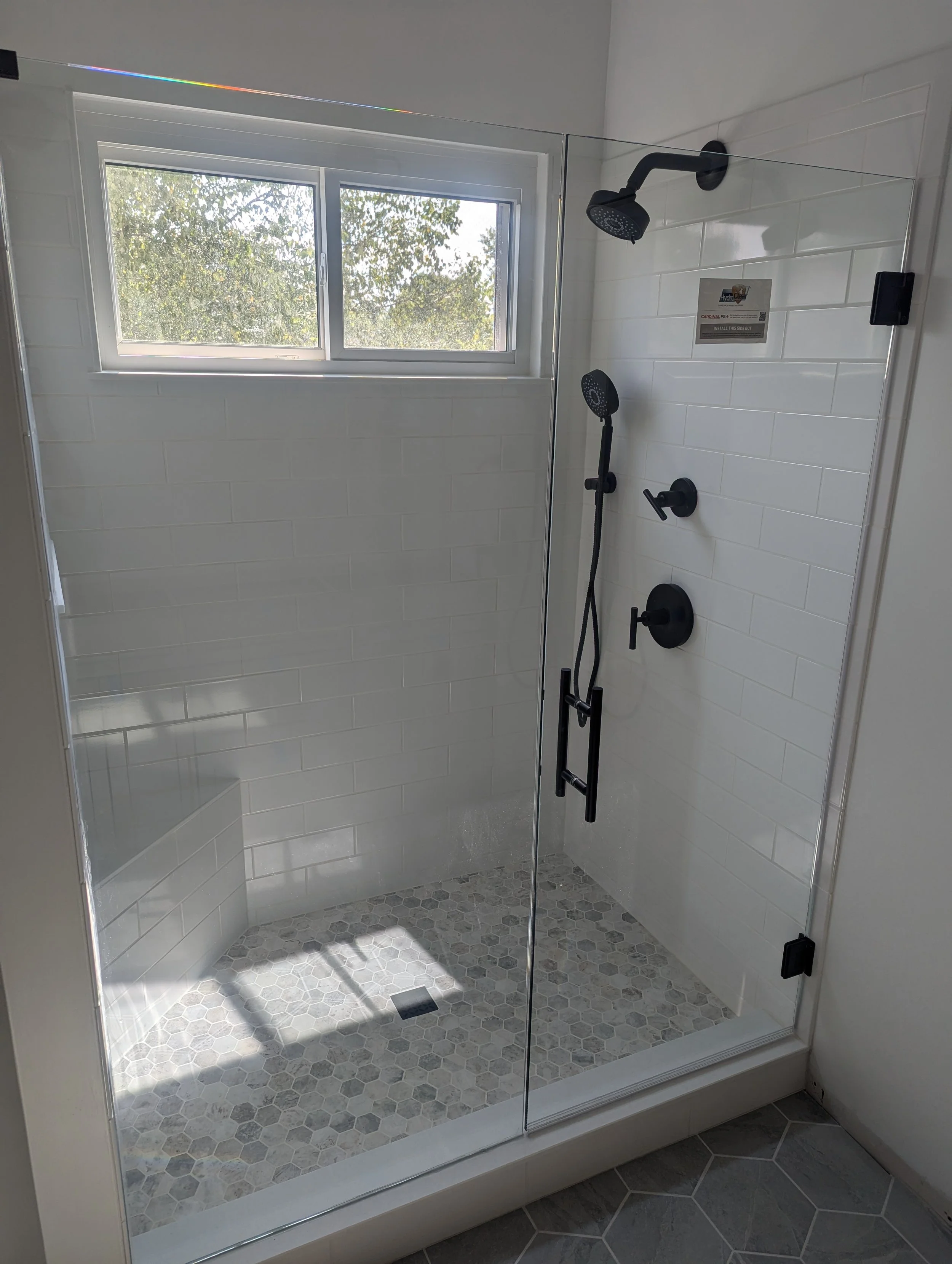 Modern shower with black fixtures, glass door, white subway tile walls, hexagonal stone shower floor, and a window showing trees outside.