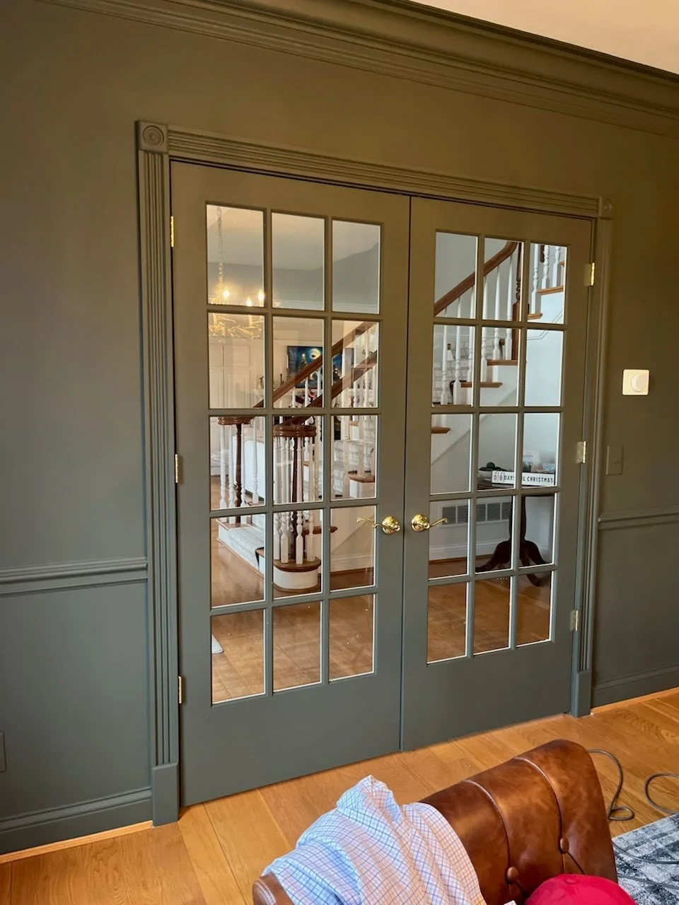 Behind double glass-paneled doors with brass handles, a view into a home's interior reveals a staircase with wooden steps and white balusters, a hallway, and a small side table with decor, all with hardwood floors and elegant trim.