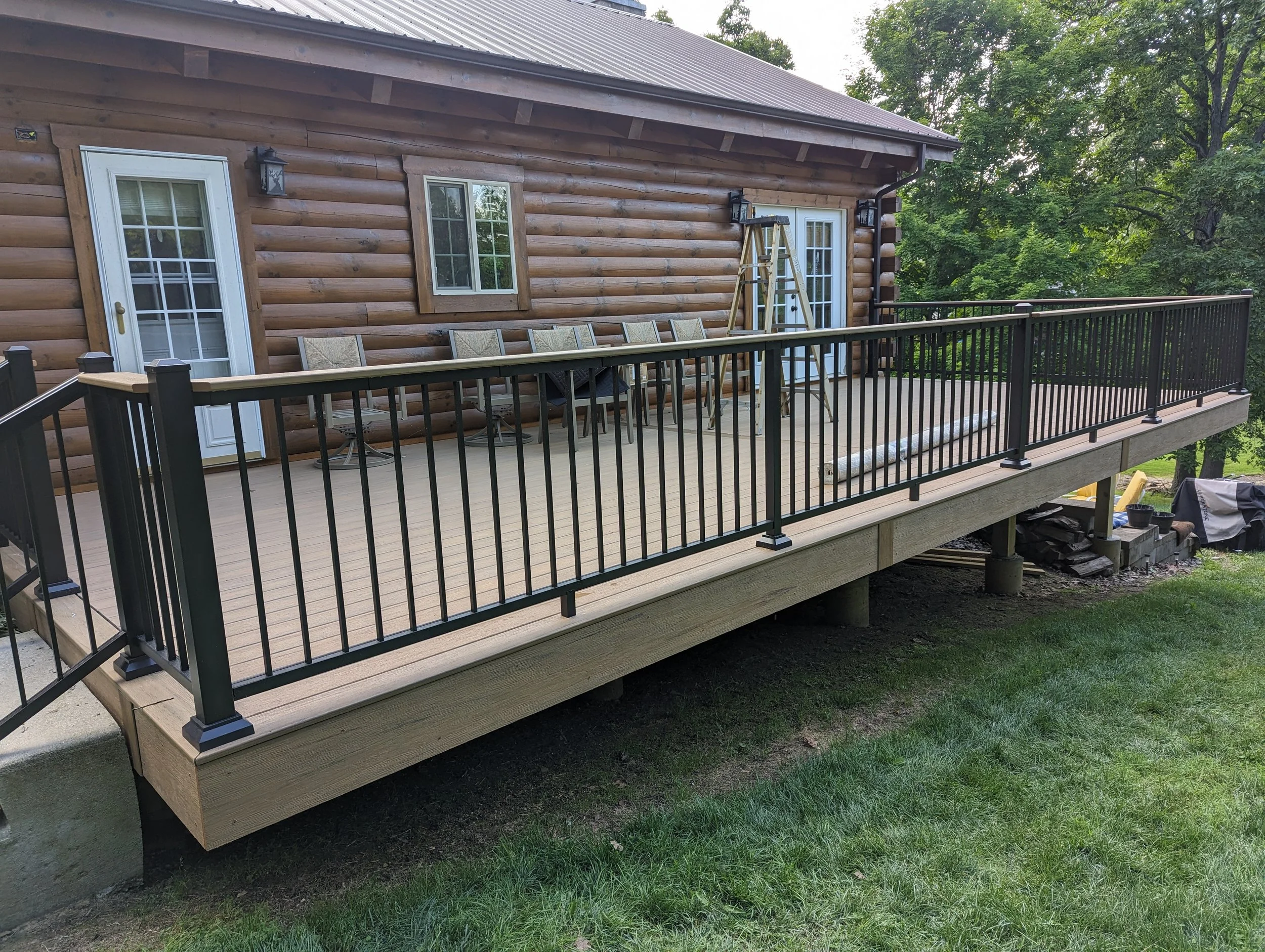 Newly constructed wooden deck with black metal railing attached to a log cabin-style house, with outdoor chairs, a small table, and a ladder leaning against the house.