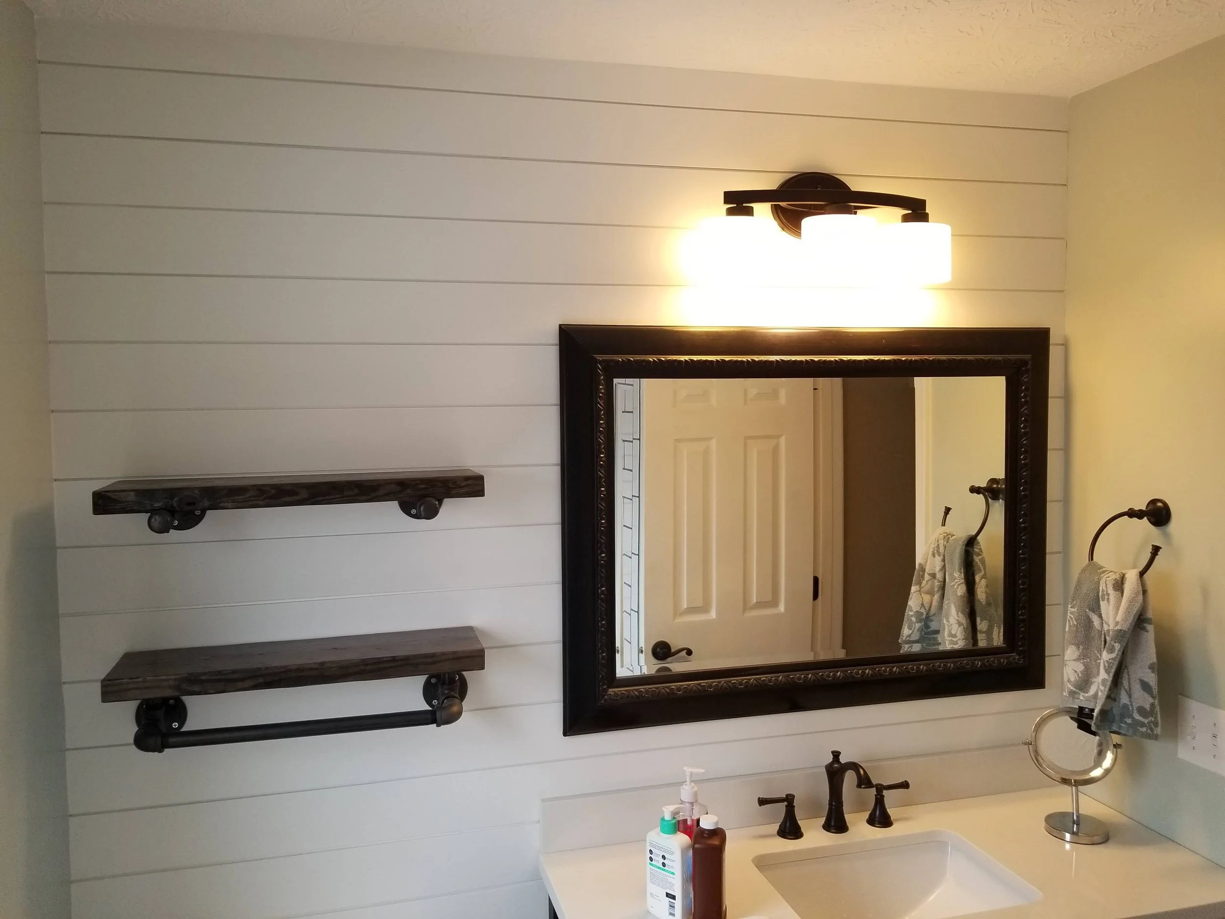 Bathroom vanity with a large mirror, two shelves, and a wall-mounted light fixture. The countertop has soap dispensers and a small mirror, with one towel hanging from a ring on the wall.