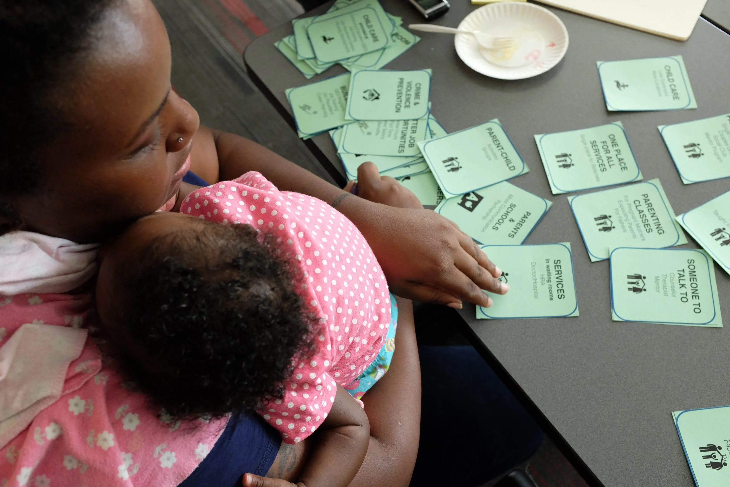 A woman and child sitting at a table with various green and blue informational cards about services and parenting. This was a participatory design activity called "card sort" used to identify which services and goals were important to that parent.