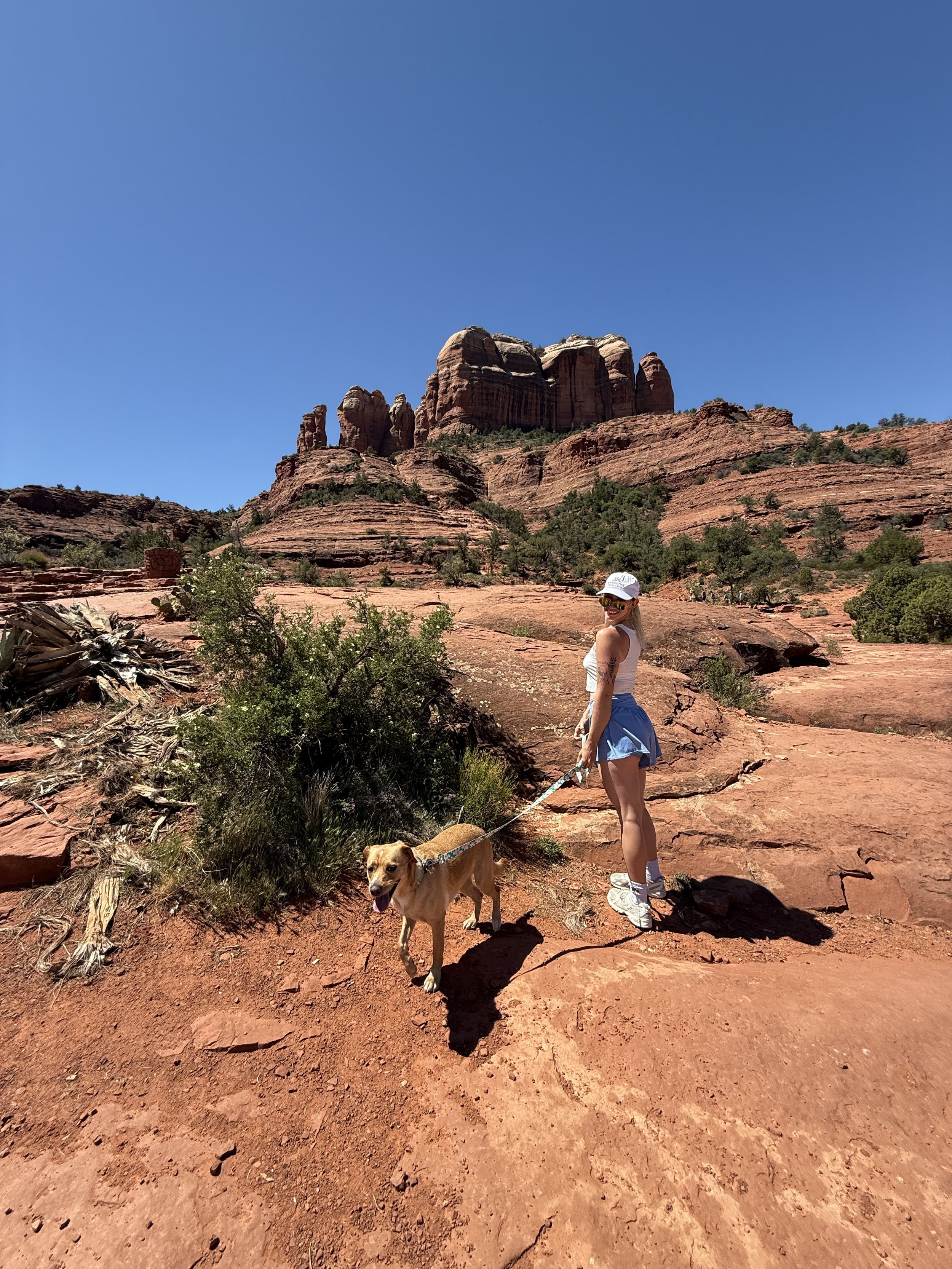 A woman and her dog hiking on a red rocky trail in a desert landscape with red rock formations and a clear blue sky.