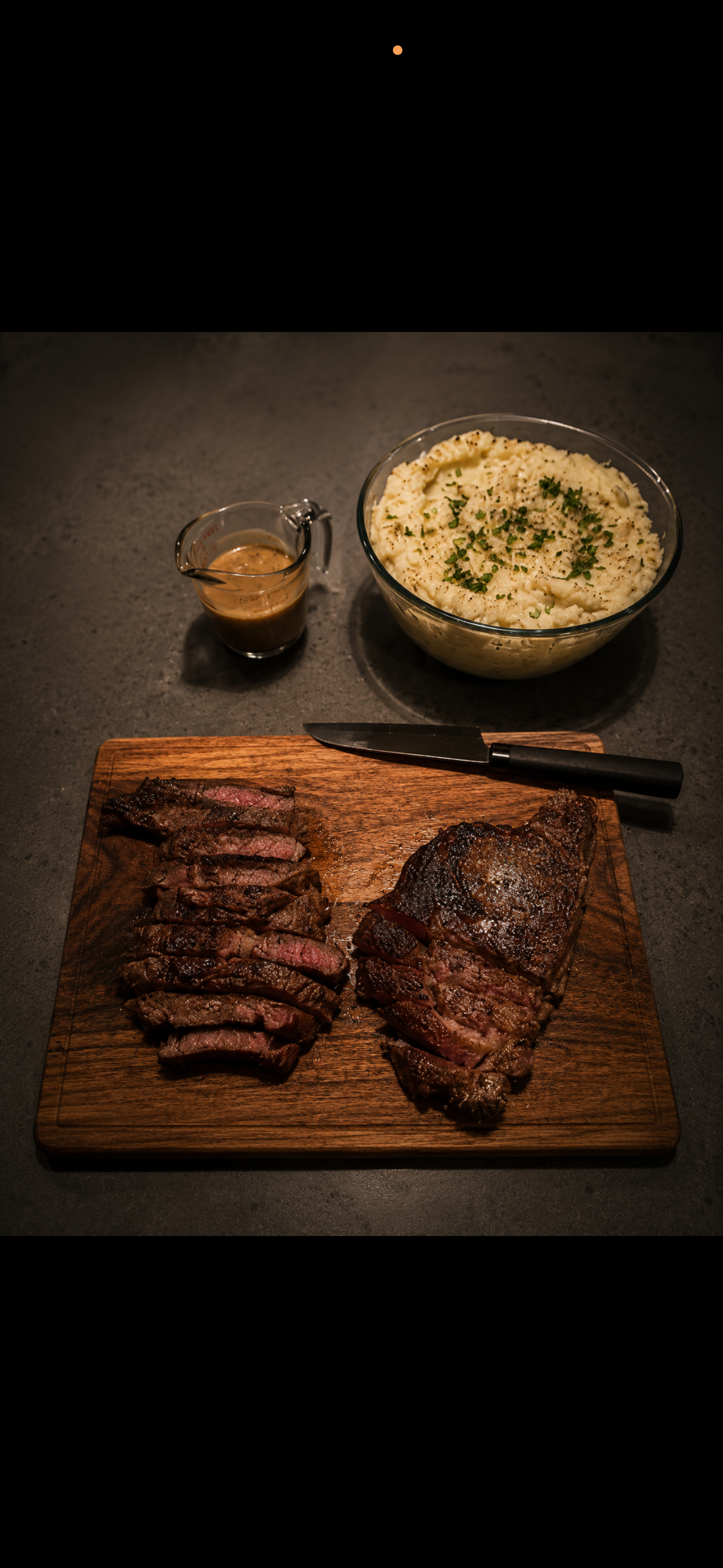 Sliced cooked steak on a wooden cutting board, with a large cooked steak and a knife. In the background, there is a bowl of mashed potatoes garnished with herbs, a small pitcher of gravy, and a dark surface.