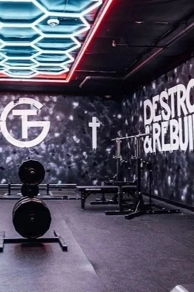 Empty weightlifting gym with black walls, illuminated ceiling hexagonal lights, and motivational slogans like "Destroy Greed" on the wall.