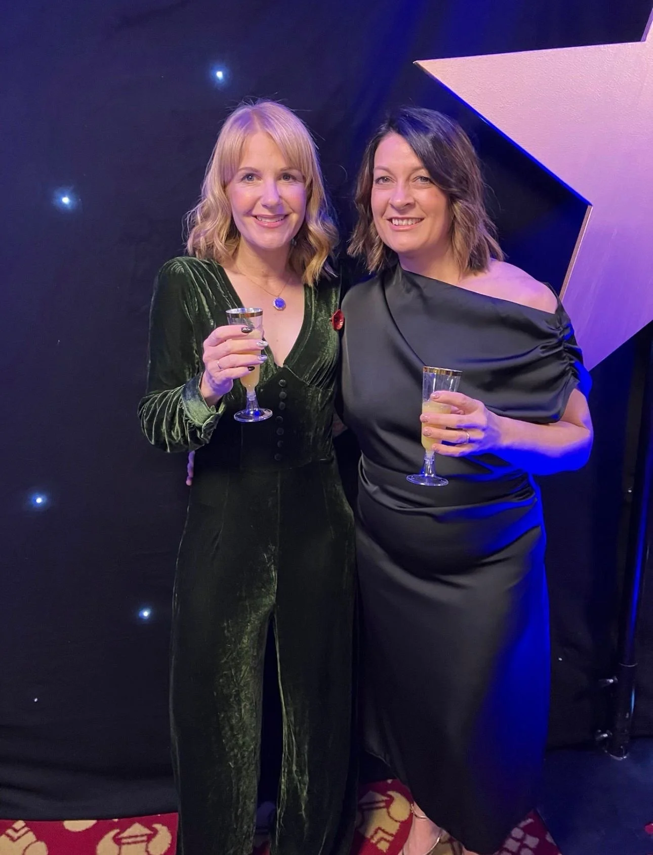 Two women in formal attire holding champagne glasses at a celebration or event, standing in front of a black backdrop with small lights and a large star-shaped decoration.