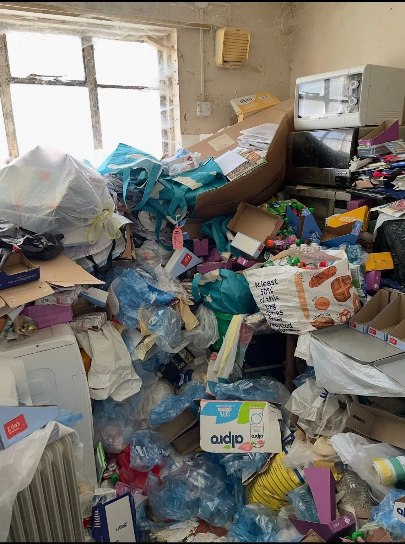 A cluttered room with scattered plastic bags, cardboard, and miscellaneous trash. A small microwave is on a makeshift table next to a computer monitor, with a window in the background.