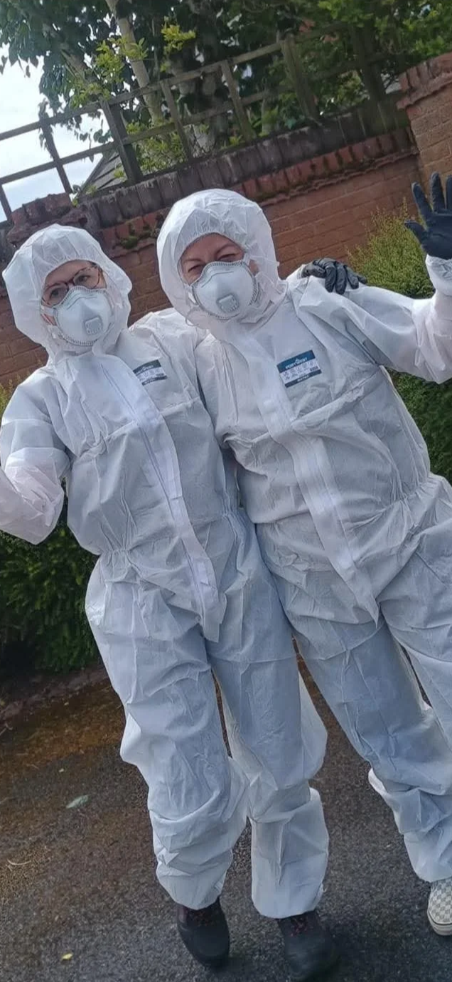 Two people wearing full protective hazmat suits, face masks, and goggles, standing outdoors in front of a brick wall and greenery, waving at the camera.