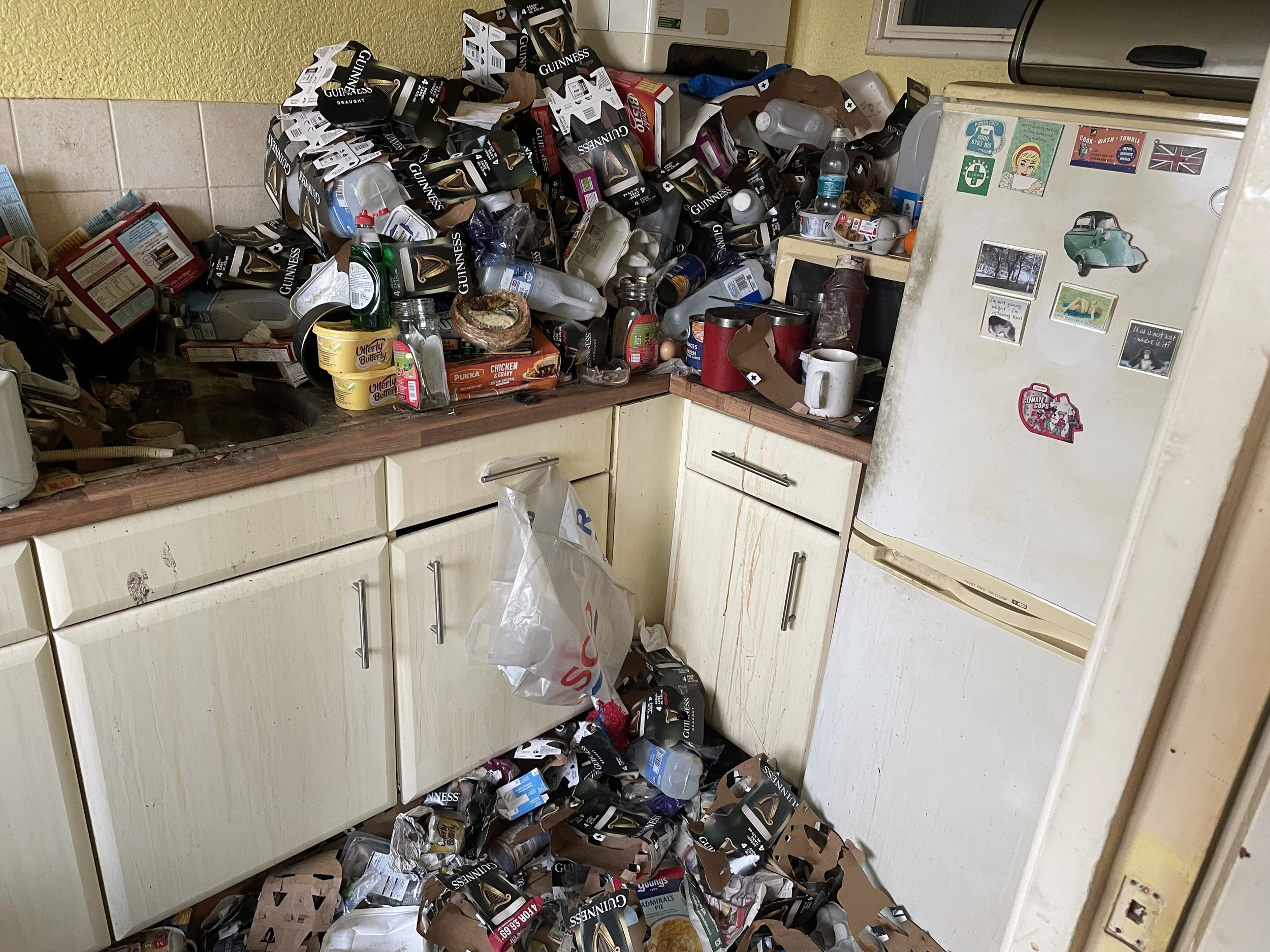 A cluttered kitchen with a messy countertop and floor covered in empty food and drink containers, including beer cans, water bottles, and food packages. The fridge has various magnets and stickers, and the kitchen cabinets are stained and disorganize