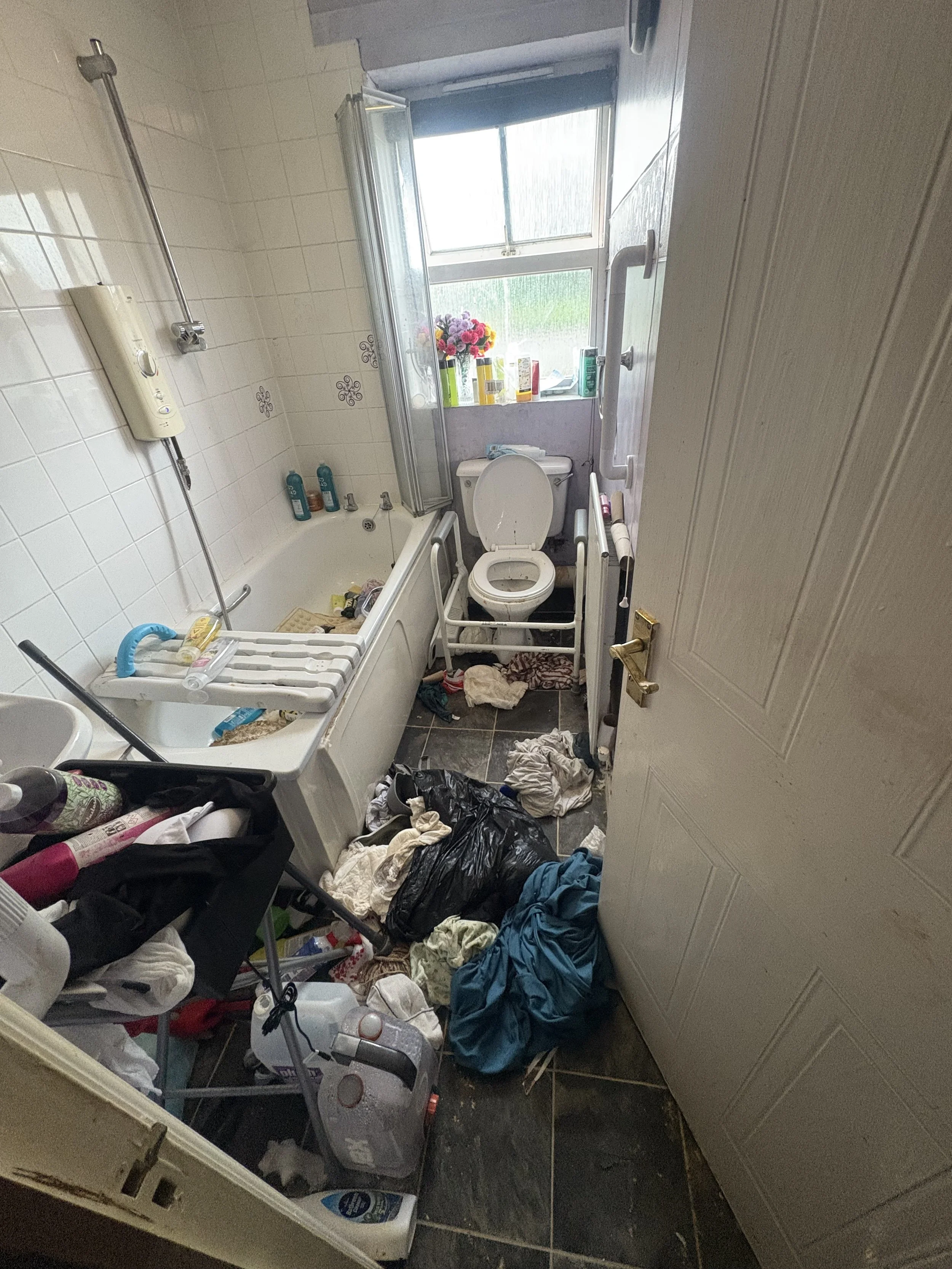 A cluttered bathroom with laundry on the floor, a bathtub filled with bottles and cleaning supplies, a toilet, and a window with flowers and toiletries on the sill.