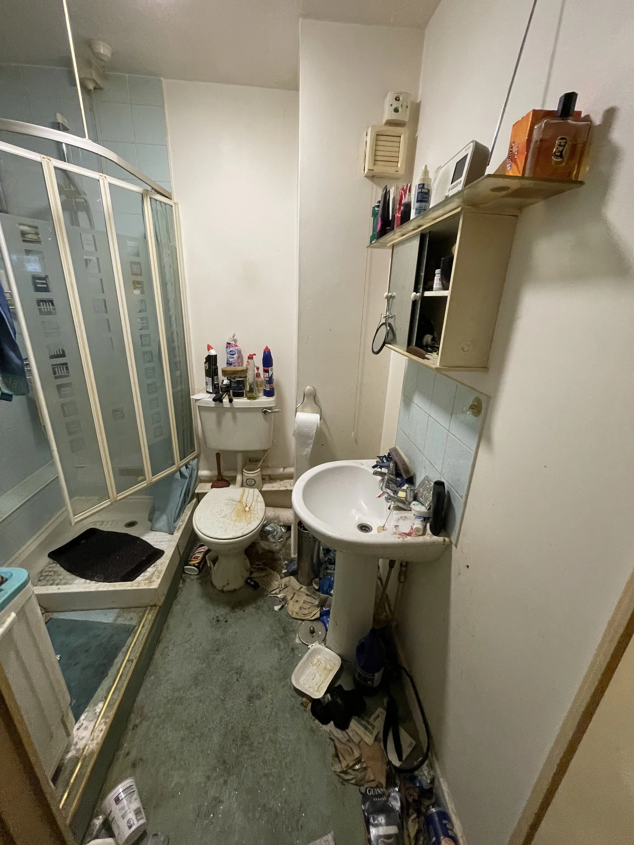 A small bathroom cluttered with trash, toiletries, and dirty items. Contains a shower with a frosted glass door, a toilet with grime, a sink with unorganized items, and a wall-mounted cabinet with personal care products.
