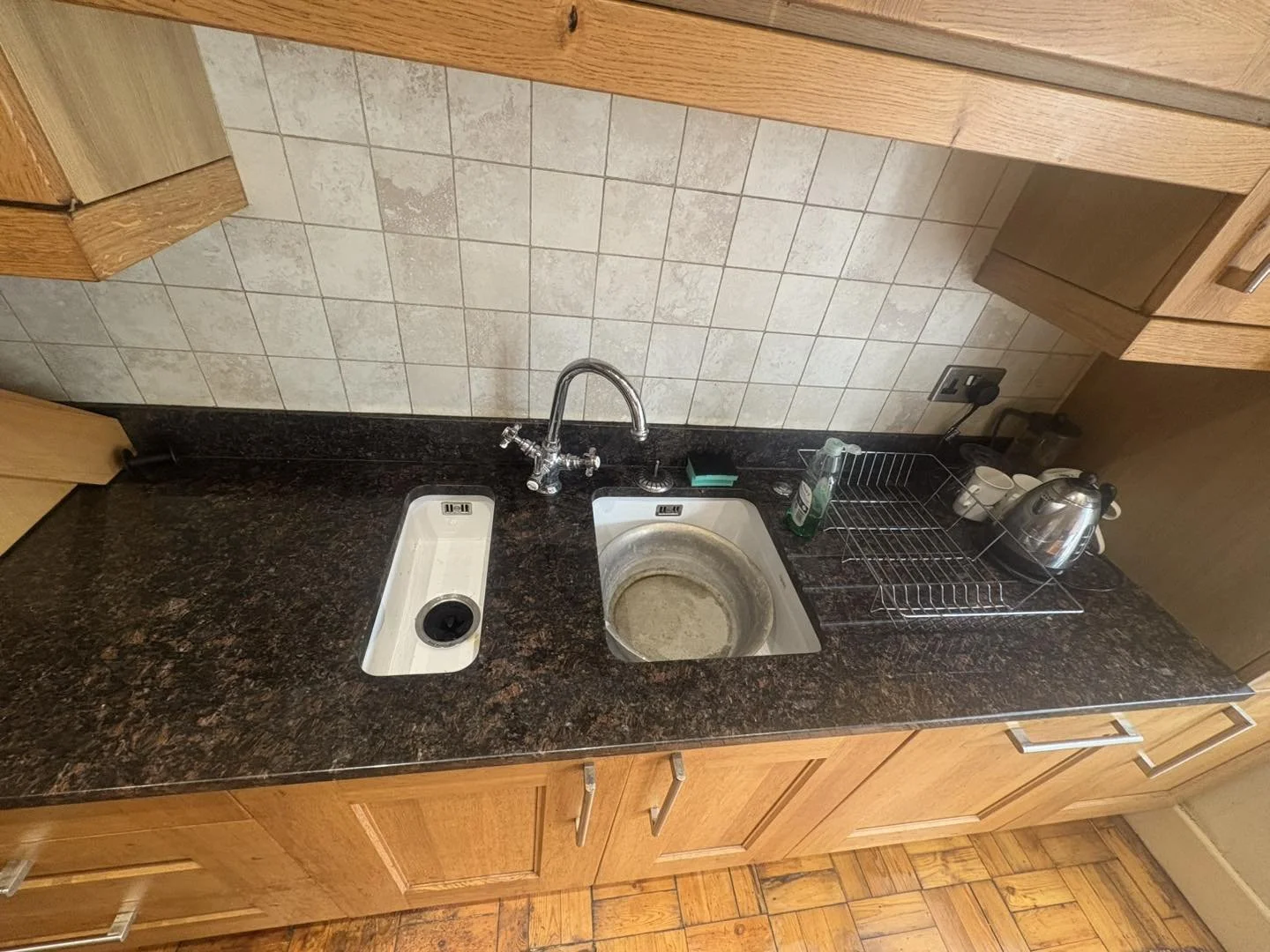 Kitchen countertop with two sinks, one with a dirty dish basin, a dish rack, a kettle, and dish soap, with wooden cabinets above and hardwood floor below.