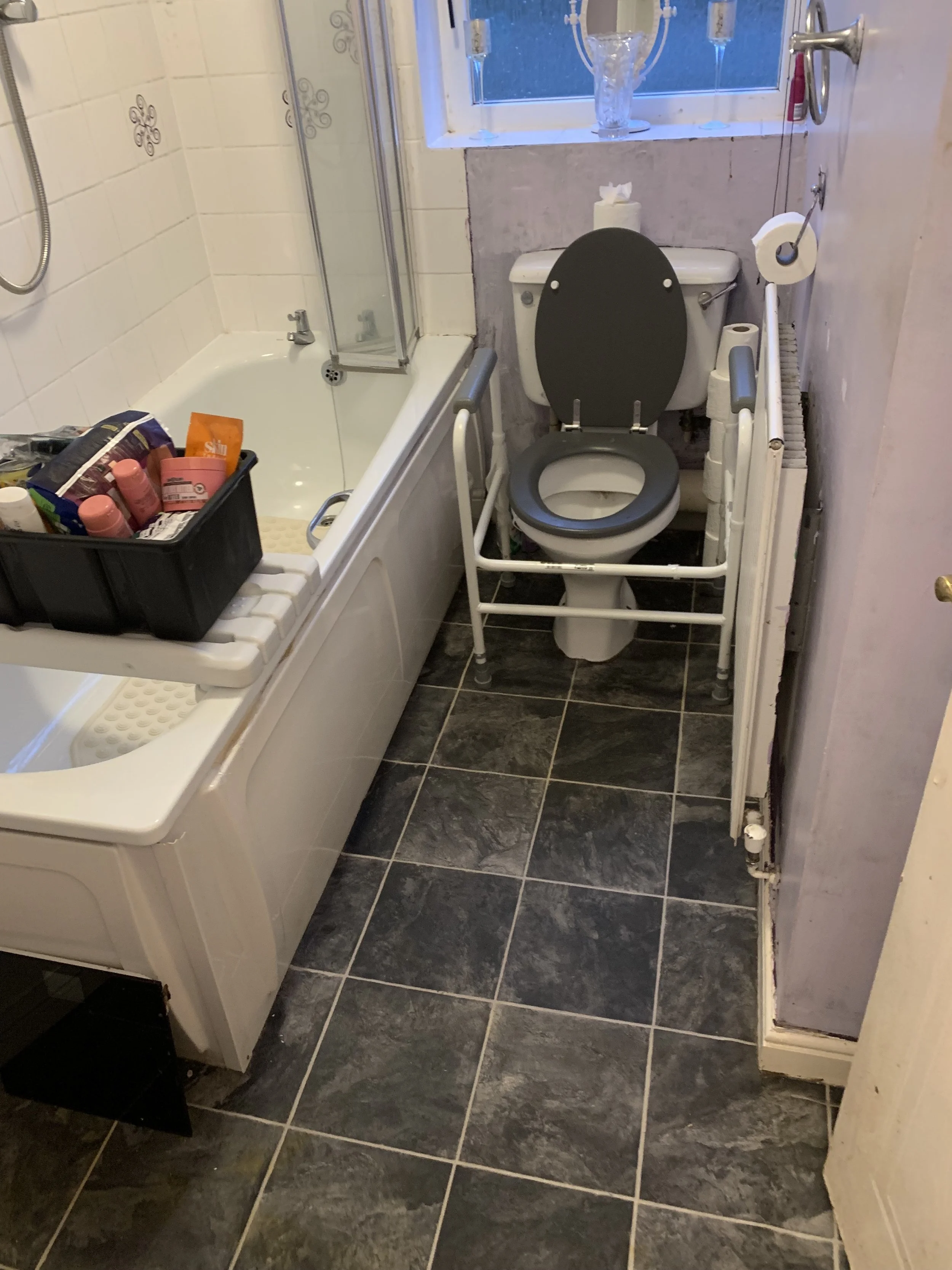 A small bathroom with a bathtub, a black storage bin with toiletries, a toilet with a medical support frame, a window, and a radiator.
