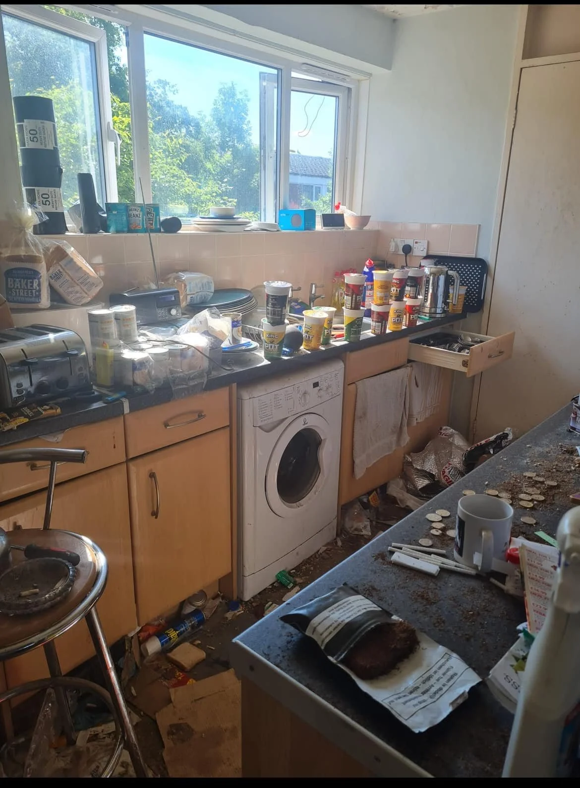 A cluttered kitchen with numerous food containers, coffee cups, and miscellaneous items scattered across the counter. The floor is dirty with trash and coins. The window shows a sunny outdoor scene with trees.
