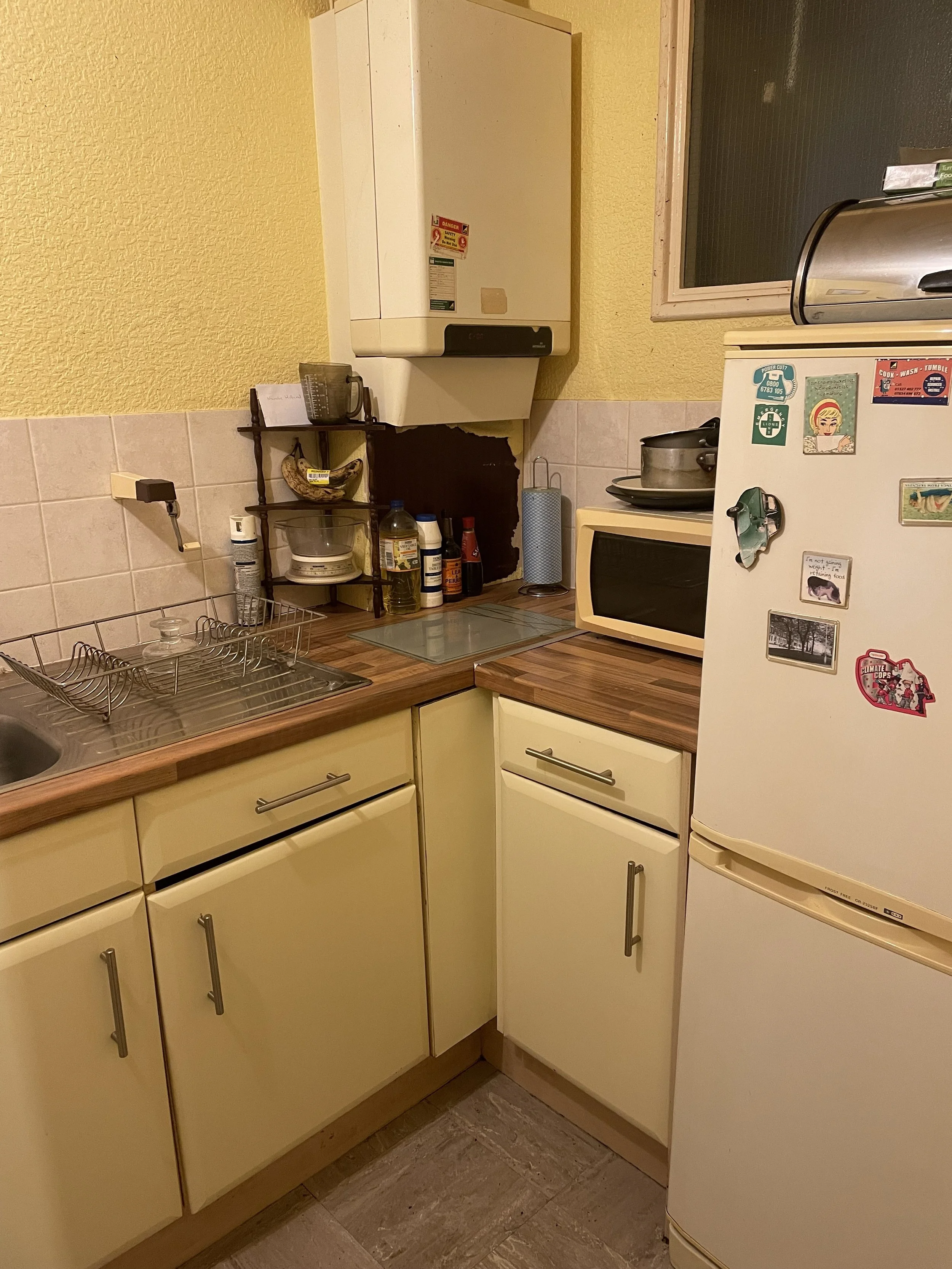 Cozy kitchen corner with yellow cabinets, a wooden countertop, a sink with a dish drying rack, a small white microwave, a white refrigerator with magnets, and various bottles and kitchen items on the counter.